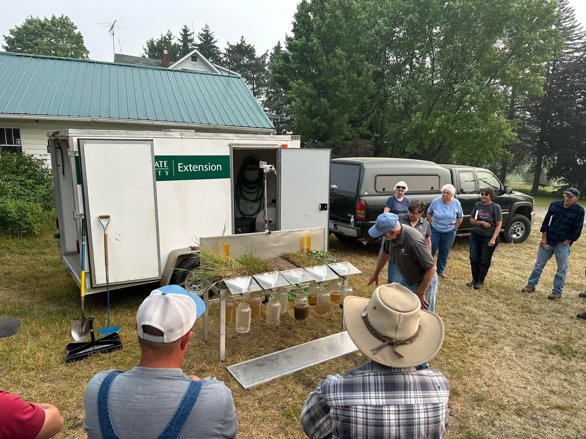 Group attending outdoor university extension demonstration near a trailer and parked truck on grassy area.