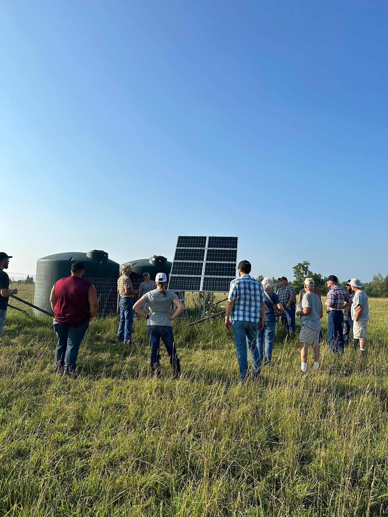 A group of people standing in a grassy field examining solar panels next to large water tanks under clear blue sky.