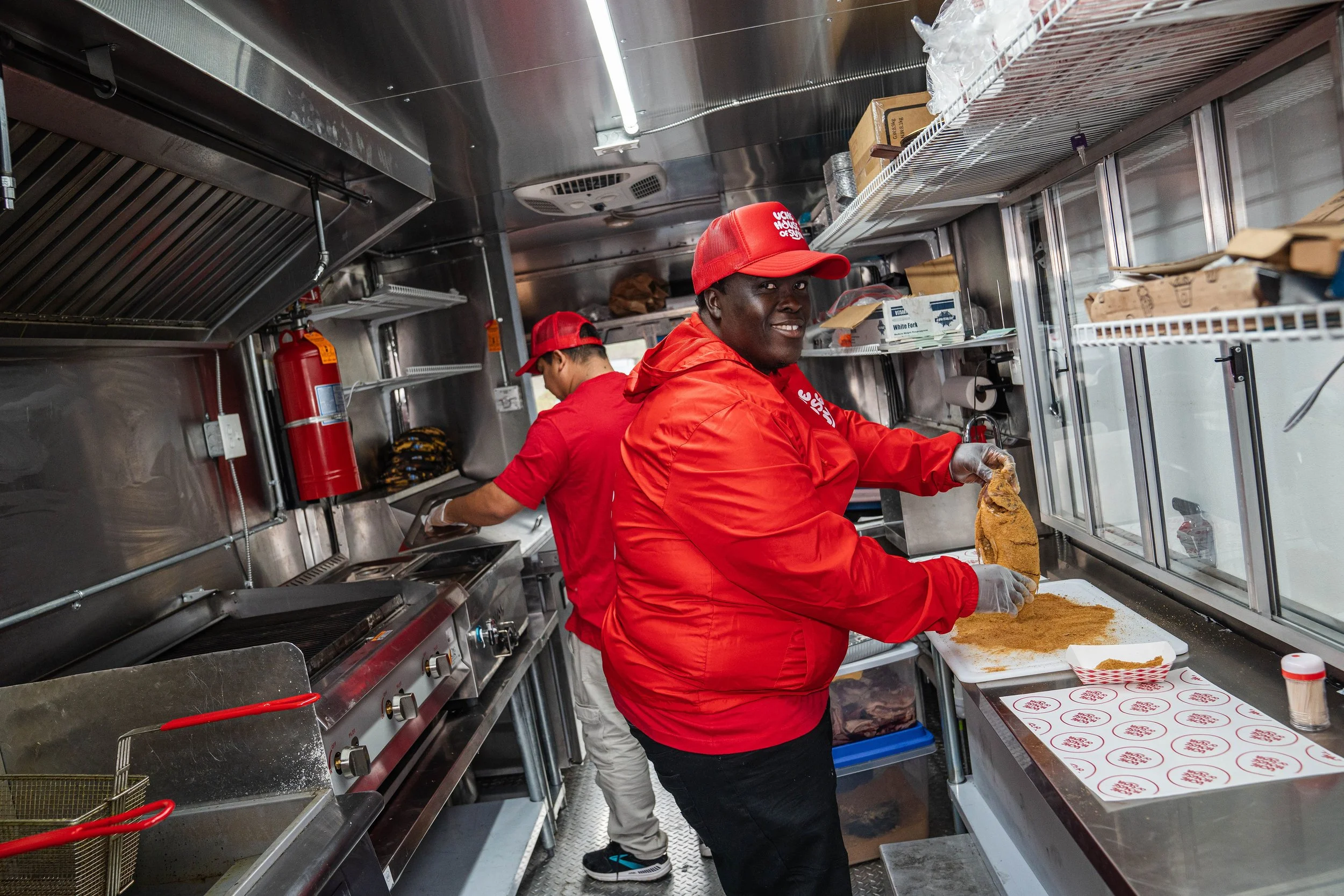 Two men cooking in a food truck kitchen, one is preparing chicken with spices and the other is working near the stove, all wearing red uniforms and caps.