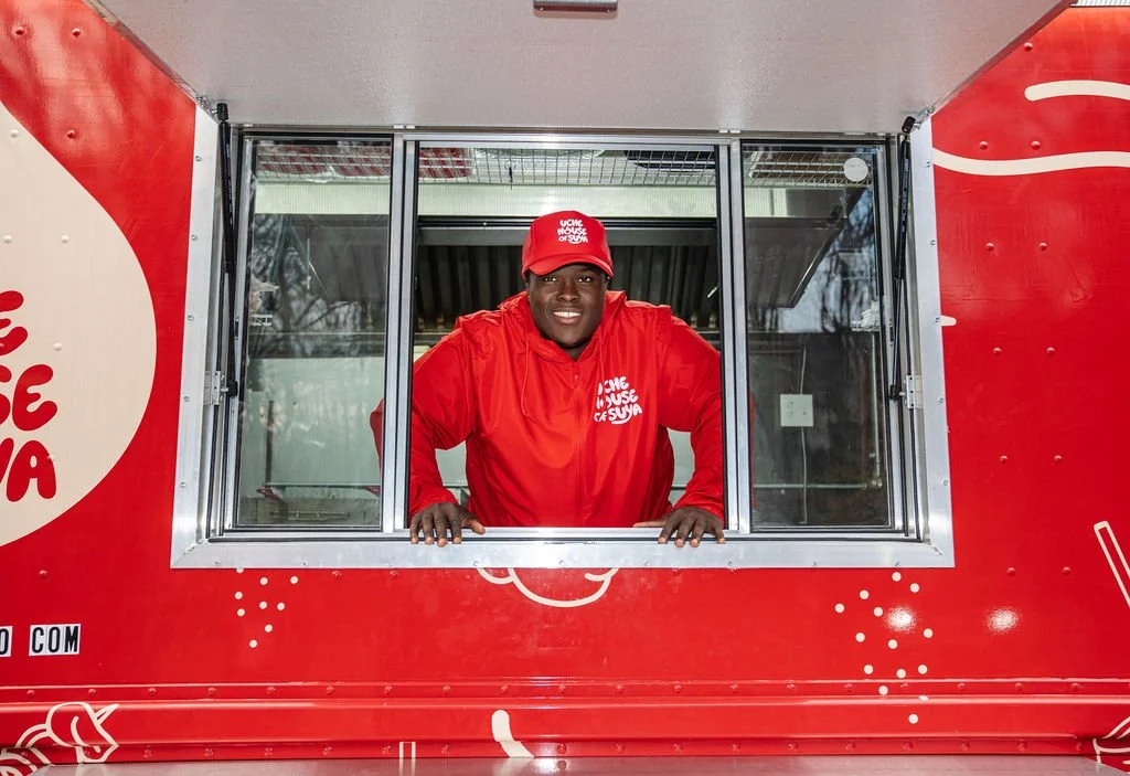 A man in red uniform and red cap smiling while leaning out of a red food truck window.