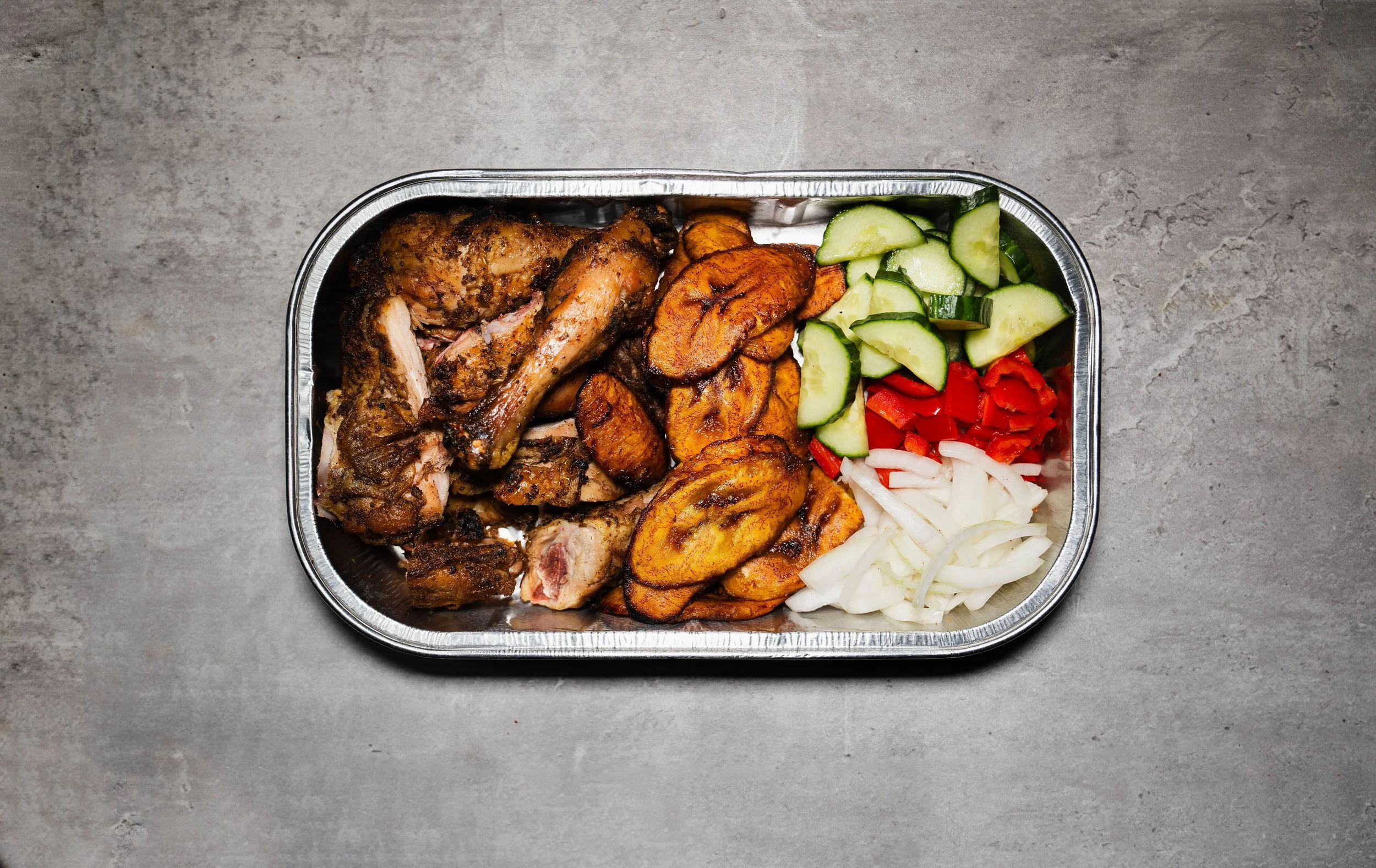 A disposable aluminum tray filled with grilled chicken, fried plantains, sliced cucumbers, chopped red bell peppers, and sliced onions, placed on a gray textured surface.