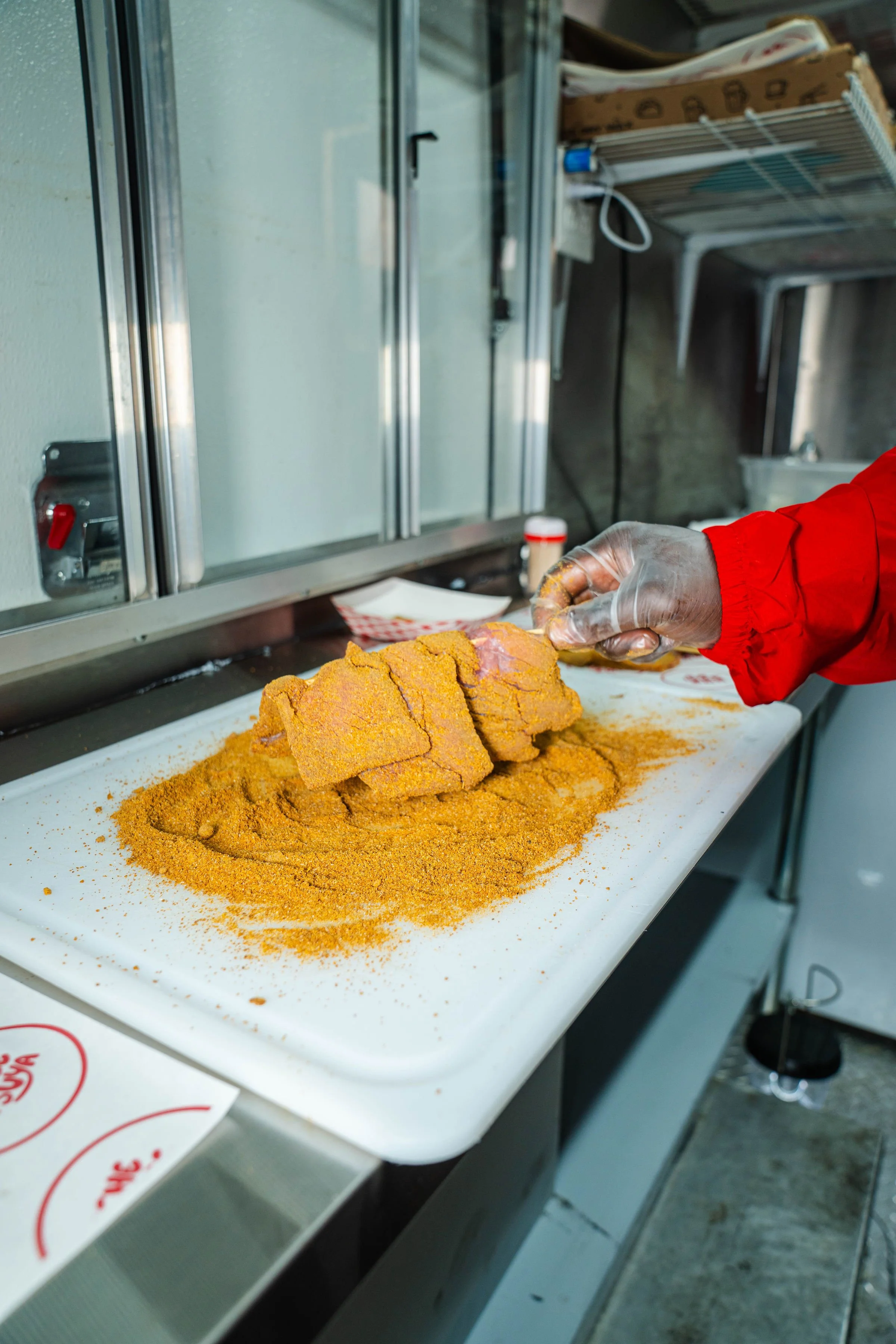 A person wearing a red sleeve and clear gloves is slicing fried fish coated in cornmeal on a white cutting board, with more pieces stacked on top, surrounded by seasoned cornmeal.