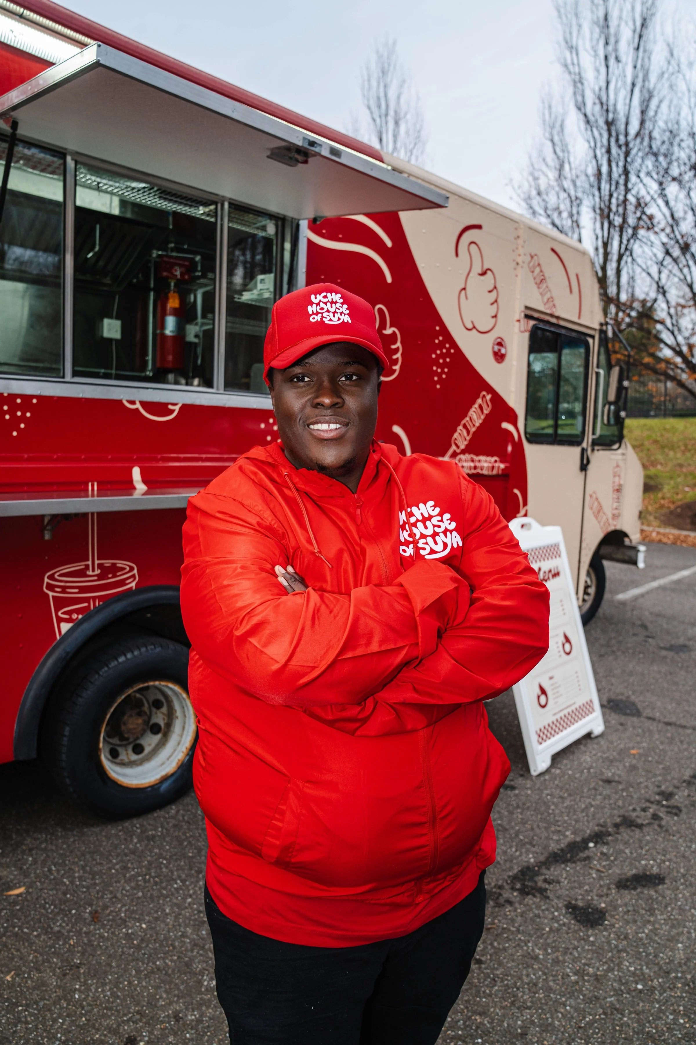 A young man in a red jacket and red cap standing with arms crossed in front of a red and white food truck with the words "The House of Suya" and illustrations of food items.