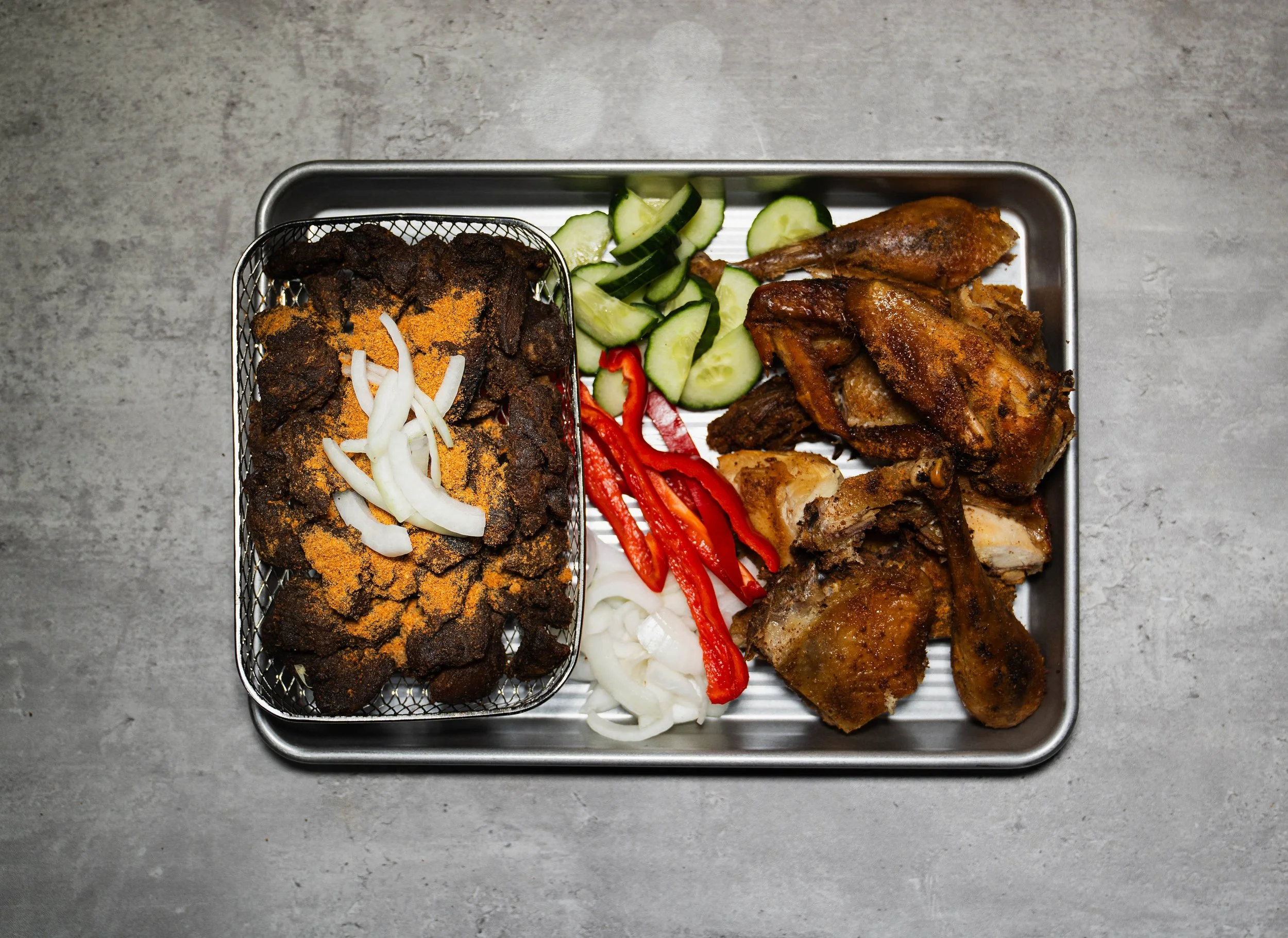 A metal tray of traditional Indonesian food including fried chicken, fried fish, sliced cucumbers, sliced onions, and red chili peppers, with a separate small basket of fried or grilled meat topped with sliced onions and seasoning.