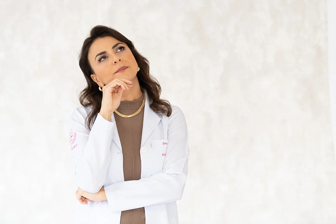 Mulher médica pensativa, vestindo jaleco branco, com expressão de reflexão, contra fundo branco.