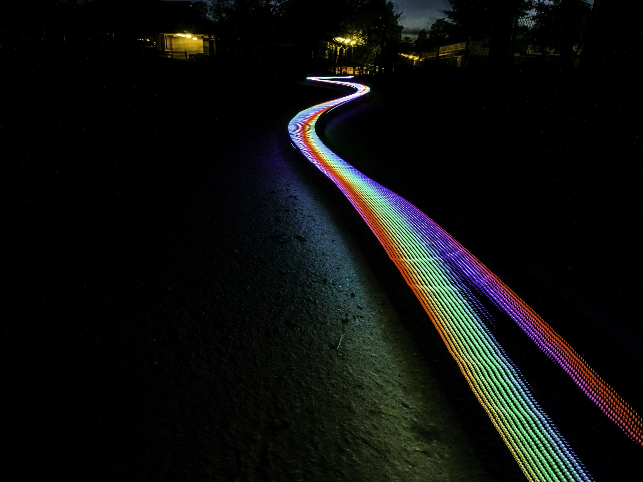 A colorful, illuminated light trail in rainbow colors, shaped like a winding path on a dark ground, with faint buildings and trees in the background at night.