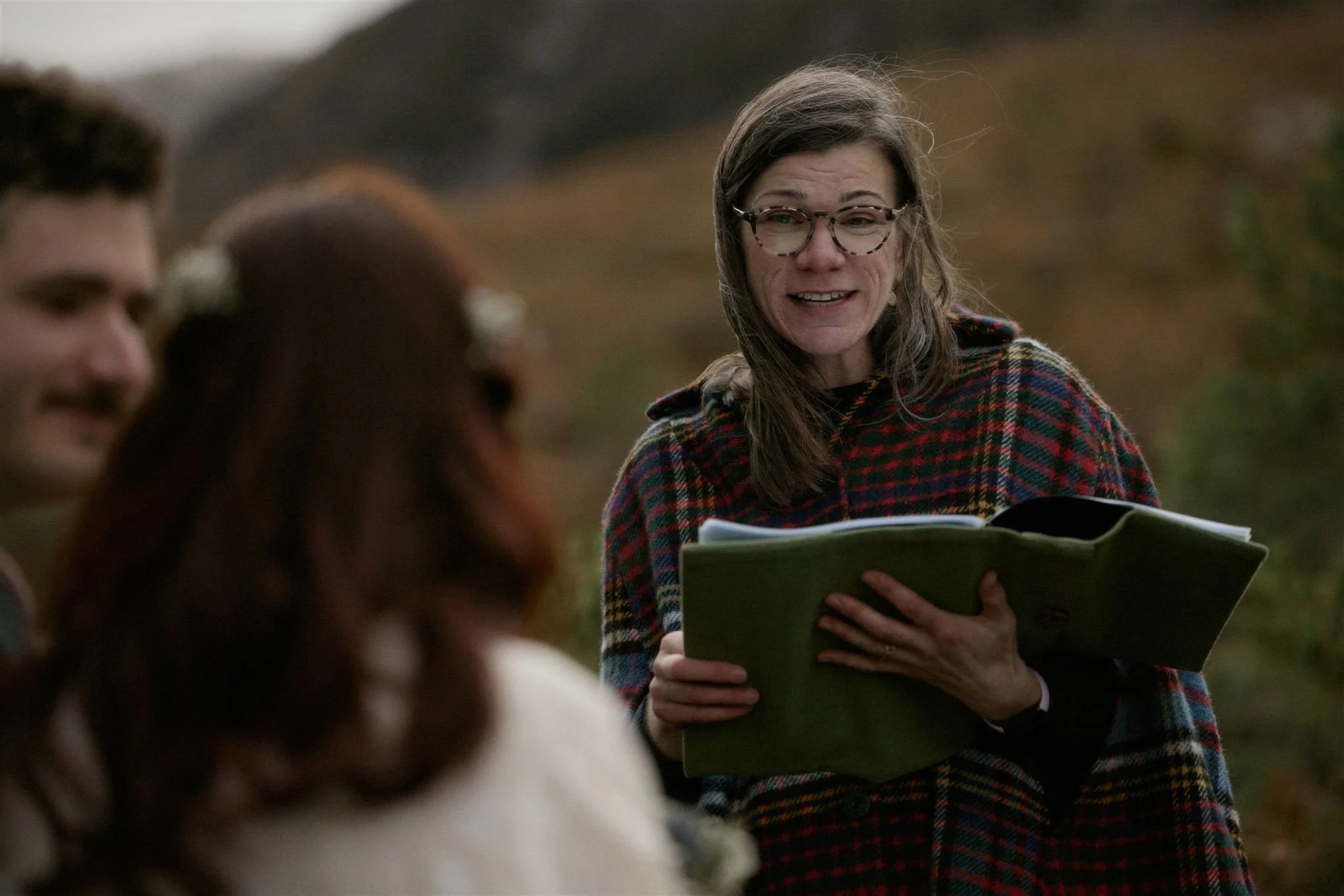 A woman wearing glasses and a colorful plaid jacket appears emotional, holding a book or folder, with three blurred people in the foreground and a blurred outdoor landscape in the background.