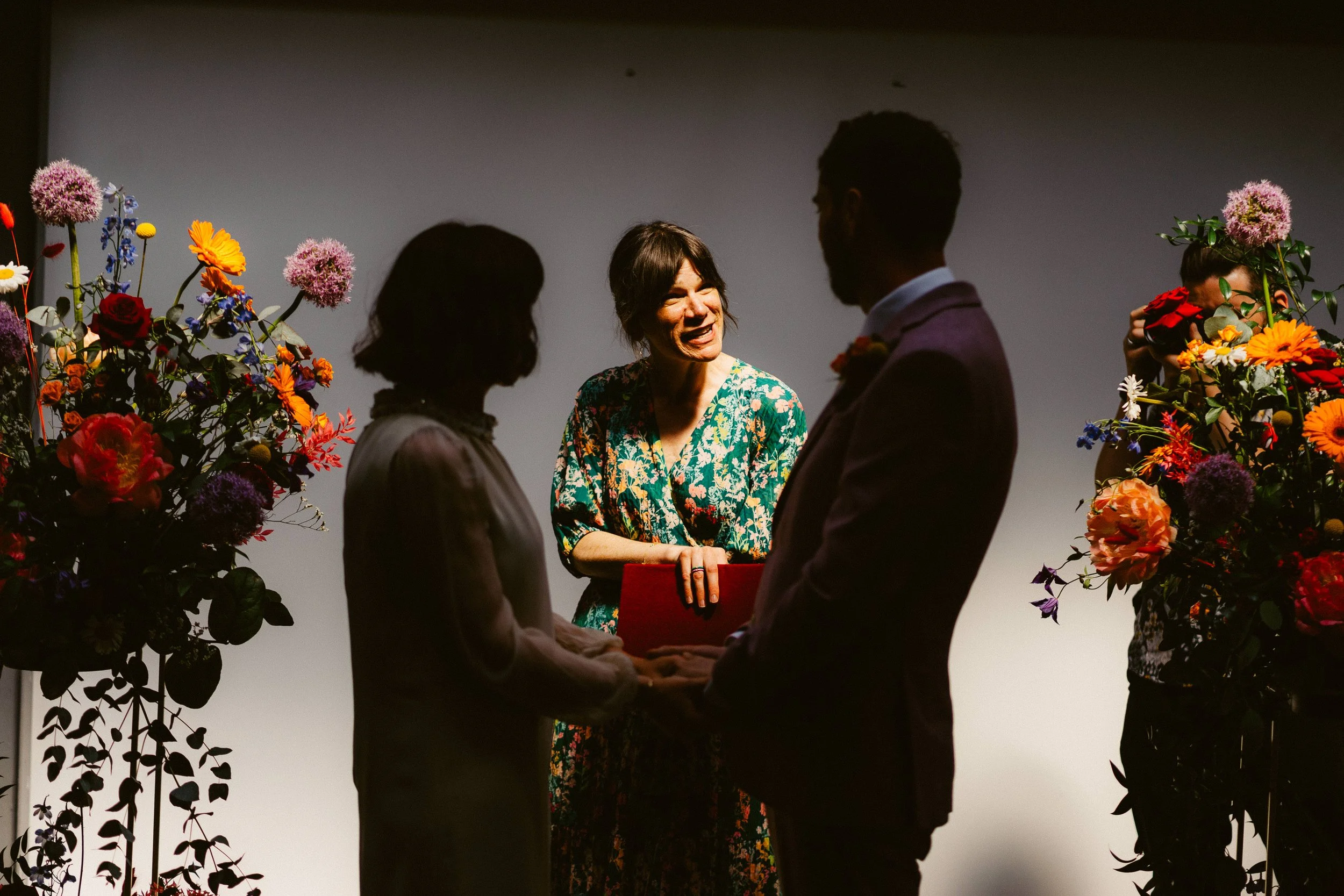 A couple holding hands during a wedding ceremony, with an officiant standing in front of them, flanked by flower arrangements.