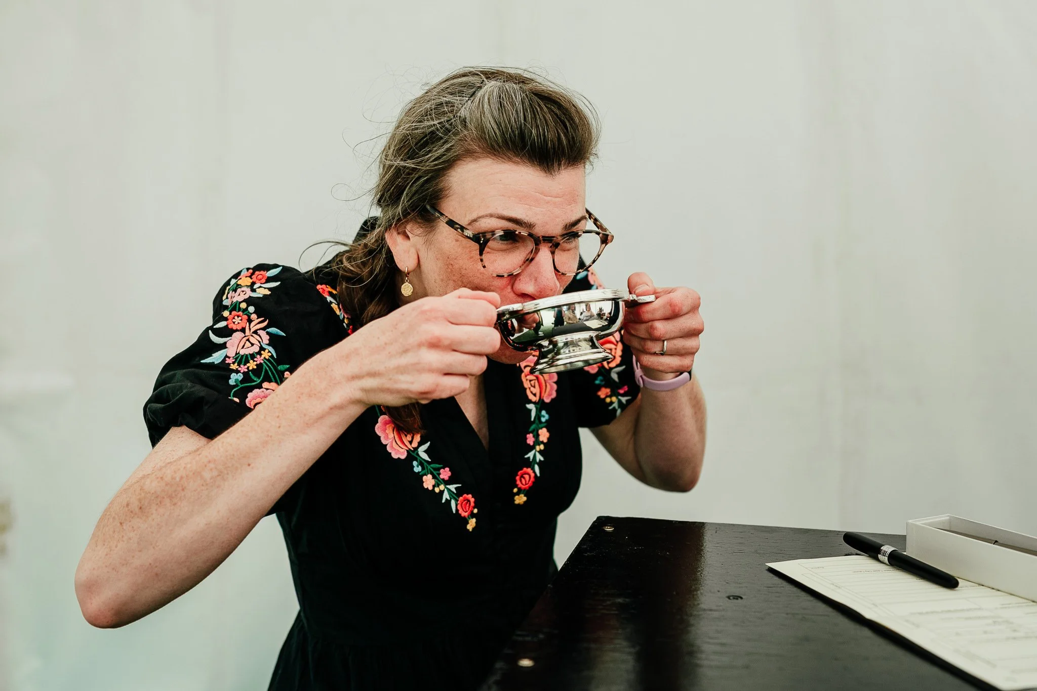 Woman with glasses and floral embroidered dress drinking from a silver cup at a desk with a notepad and pen.
