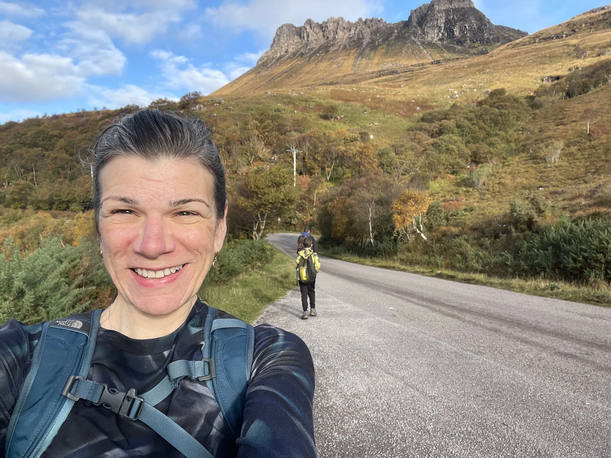 A smiling woman taking a selfie on a scenic mountain trail with two hikers in the background, surrounded by autumn colors and a mountain in the distance.