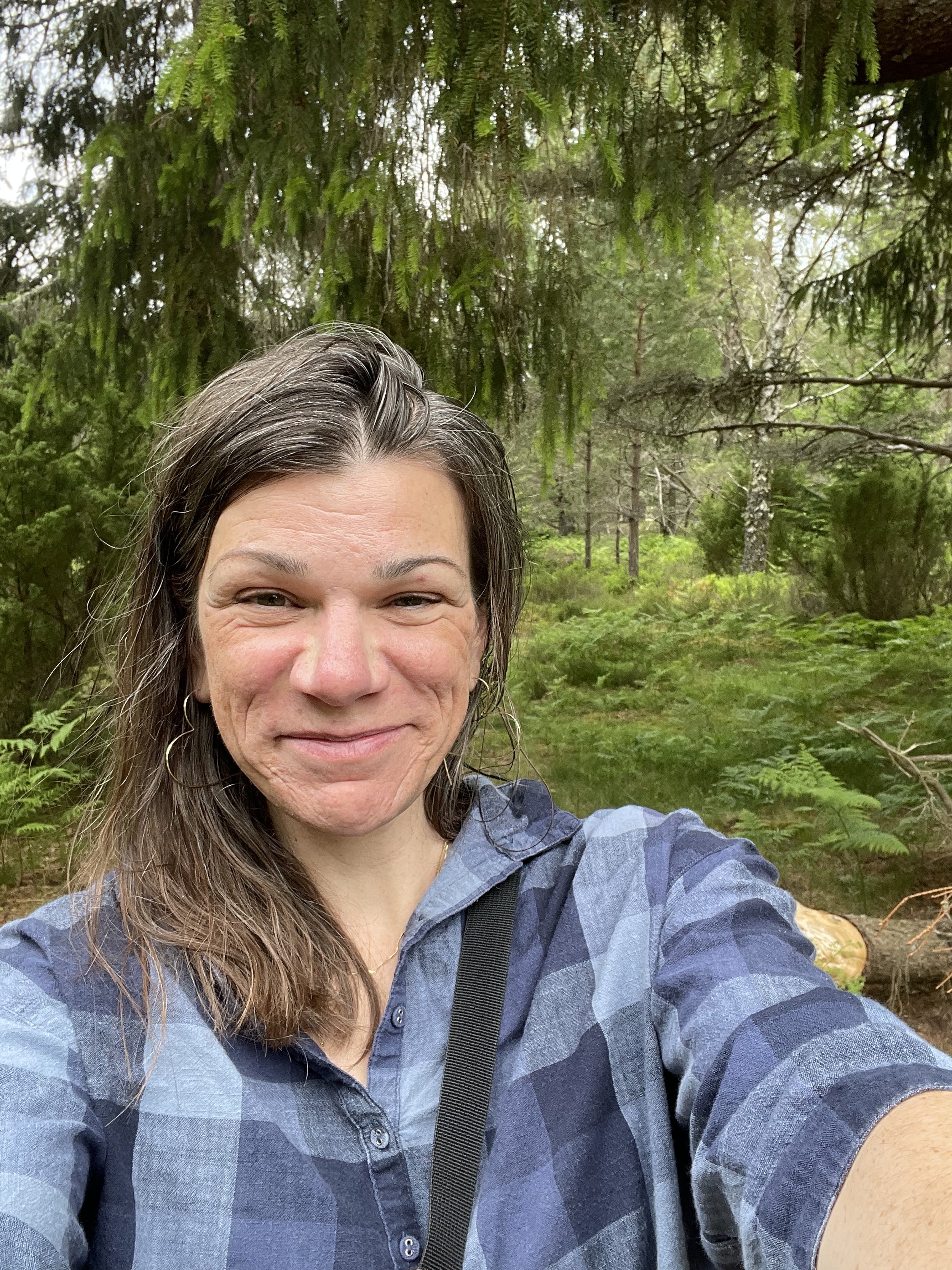 A woman taking a selfie outdoors in a lush, green forest with tall trees and ferns.