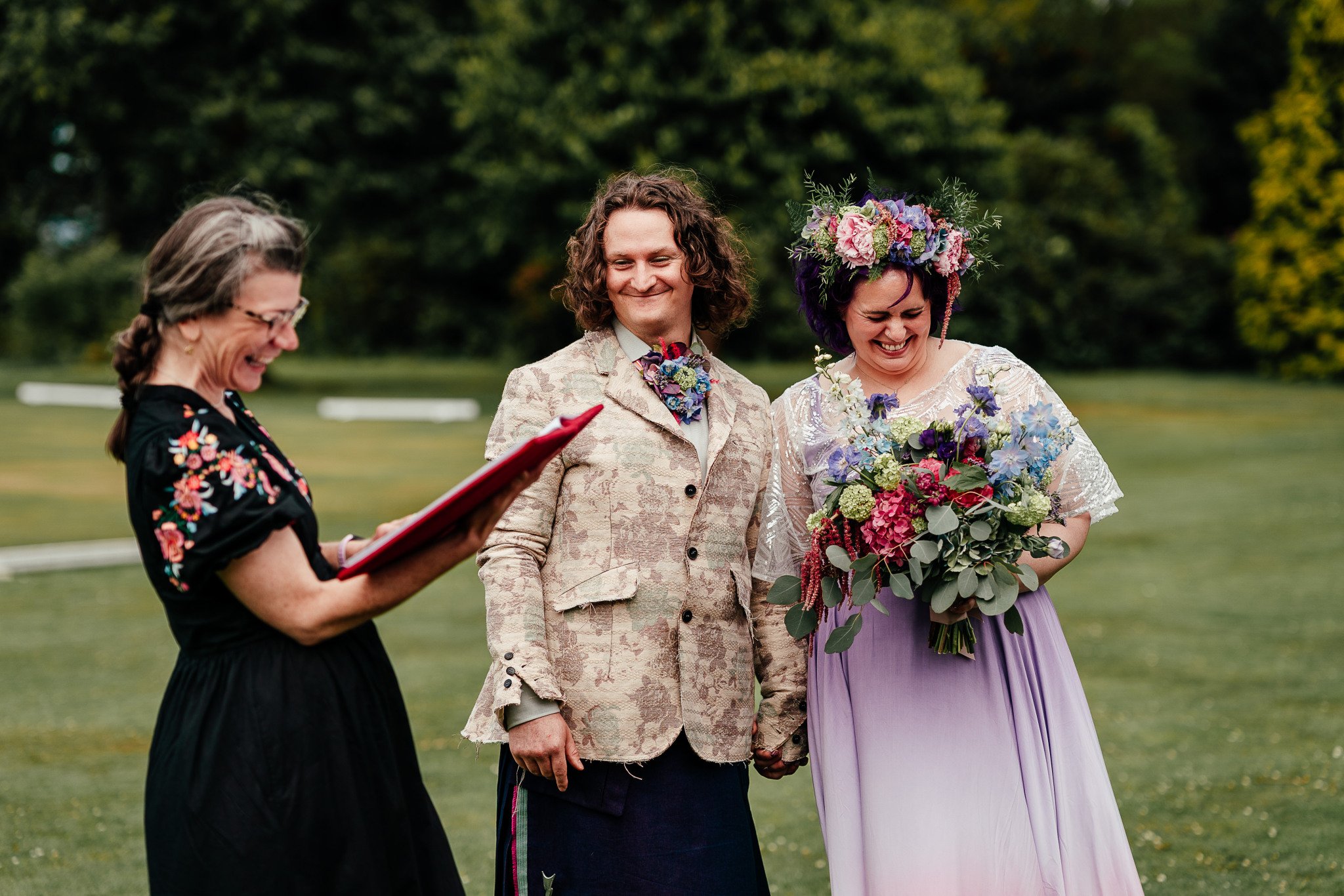 A wedding ceremony outdoors with a couple holding hands, a woman in a floral crown and a light purple dress holding a colorful bouquet, and an officiant reading from a book.
