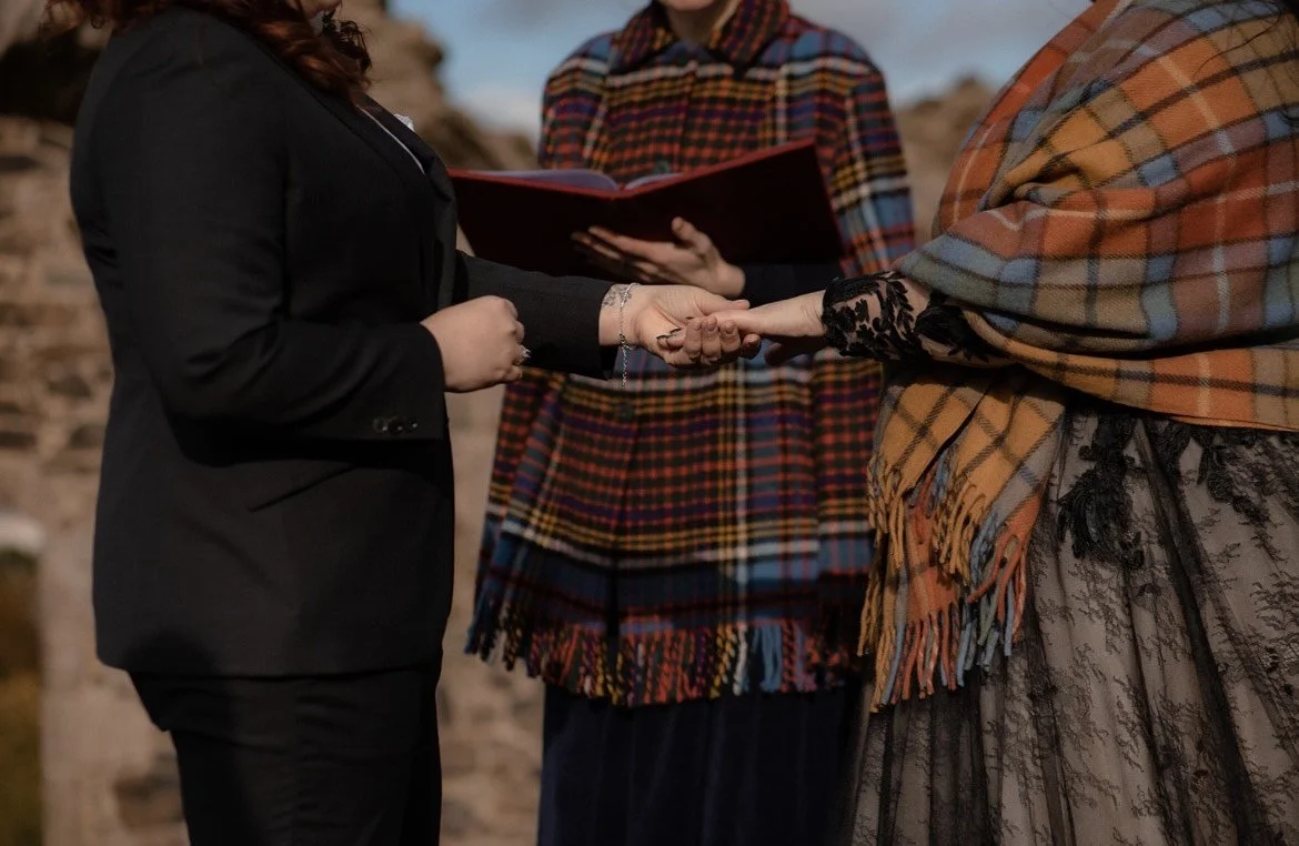 A wedding ceremony where two women are holding hands, with one woman dressed in black and the other wrapped in a plaid shawl, and an officiant standing behind them reading from a book.