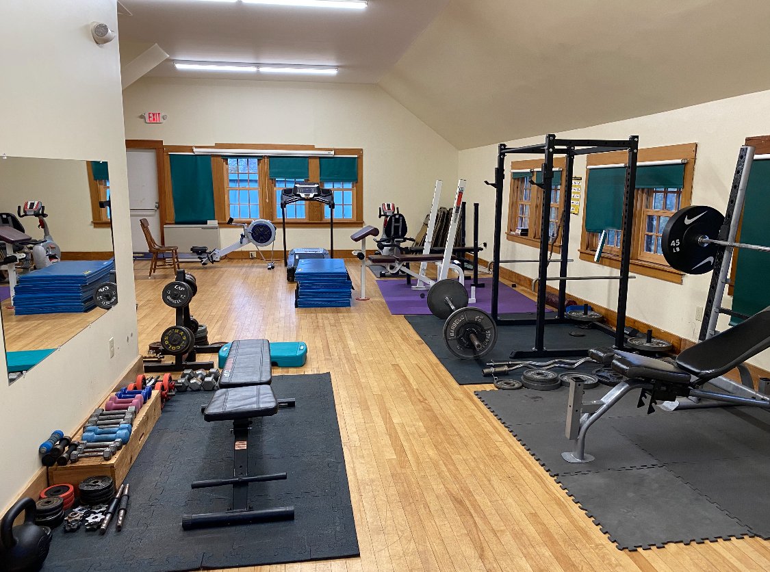 A home gym with various exercise equipment including weights, benches, squat rack, cardio machines, and stretching mats.