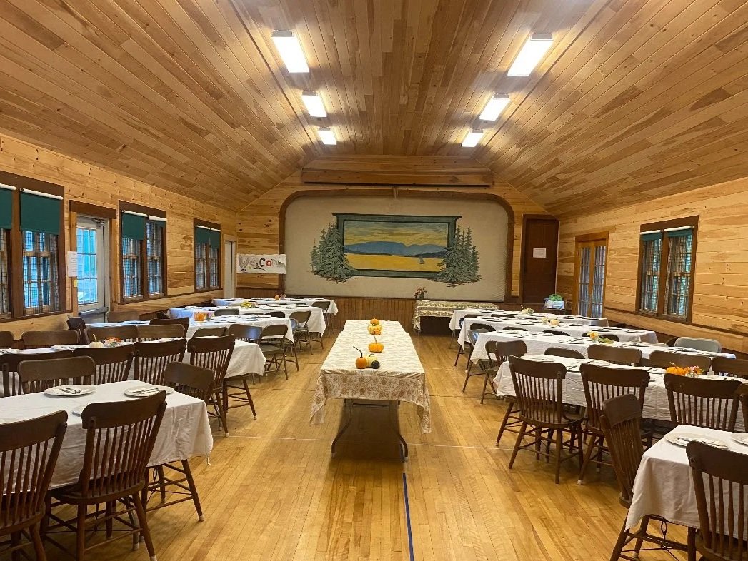 A spacious dining hall with wooden walls and ceiling, set up for an event with many round tables covered in white tablecloths, and chairs arranged around them. There are windows along both sides of the room with blue curtains, and a mural depicting a landscape at the far end.