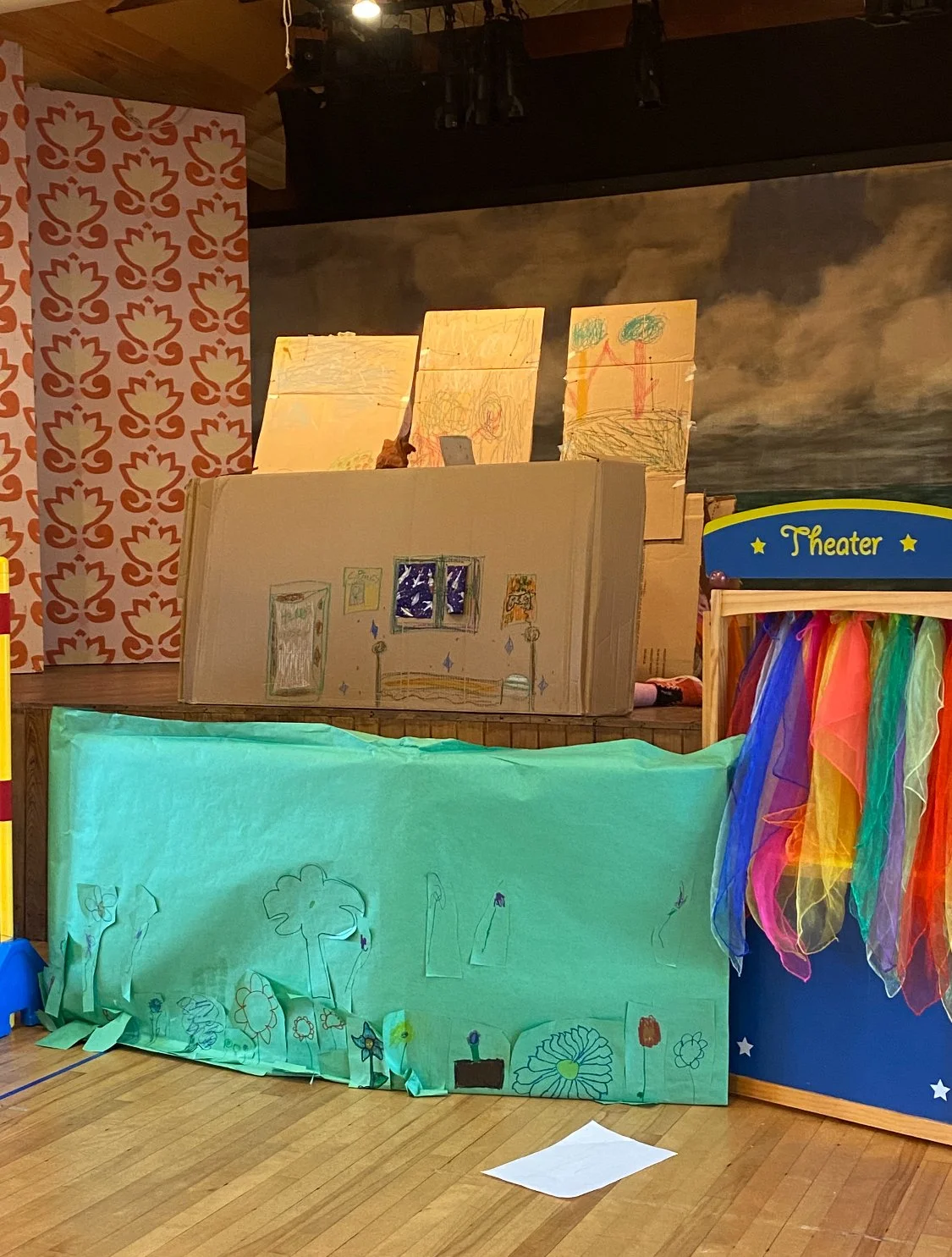 Children's artwork displayed on various colored papers, with drawings of flowers, trees, and a room with a window, set up on stage in a theater with a rainbow-colored curtain and decorative background.