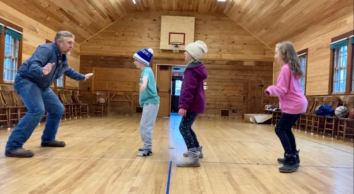 An adult man and three children in a wooden gymnasium, with the man leading a dance or jump rope activity, all standing on the wooden floor.