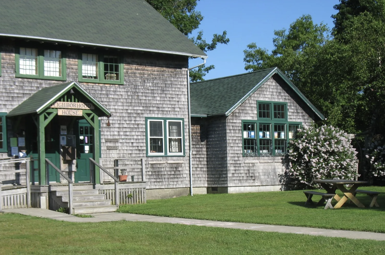 A wooden neighborhood house with a green roof and shutters, a porch with steps, a sign reading 'NEIGHBOURHOOD HOUSE,' and a surrounding yard with grass, a blooming shrub, a picnic table, and trees under a blue sky.