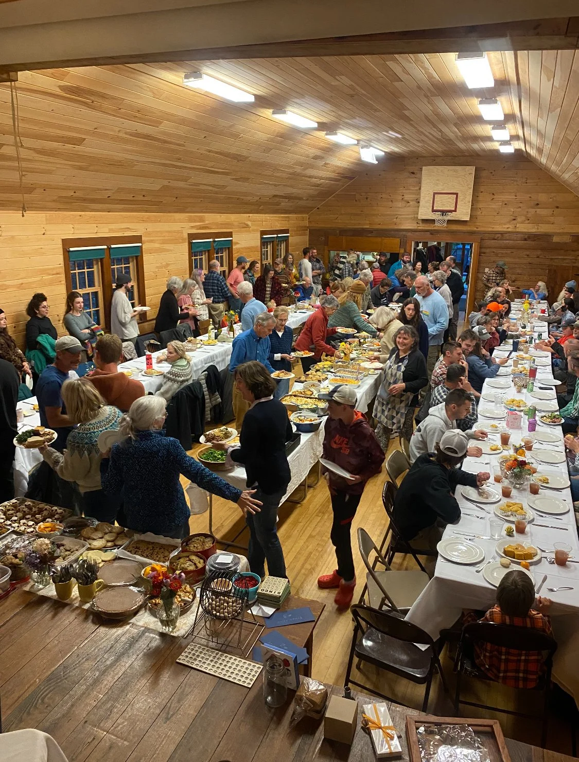 A large group of people gathered in a dining hall for a meal, with food on long tables and some people serving or eating. The hall has wooden walls and ceiling, with fluorescent lights and windows along the wall.
