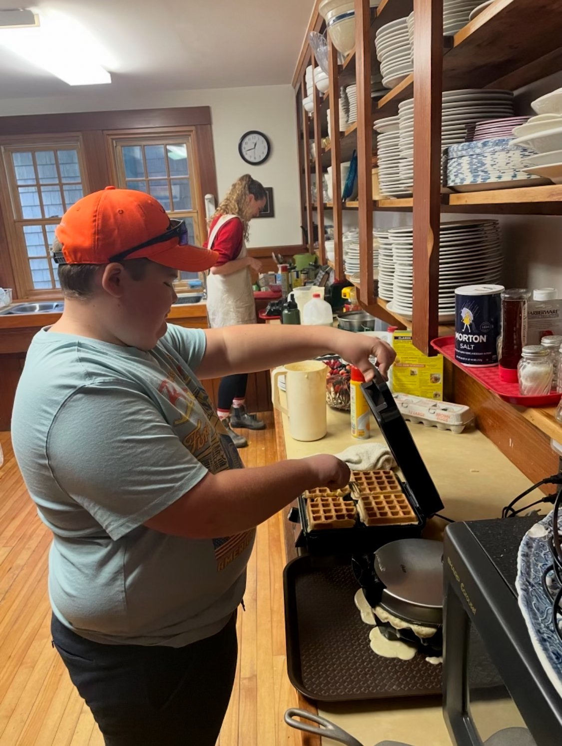 A boy with an orange cap making waffles using a waffle maker in a kitchen, with a woman in the background preparing food.