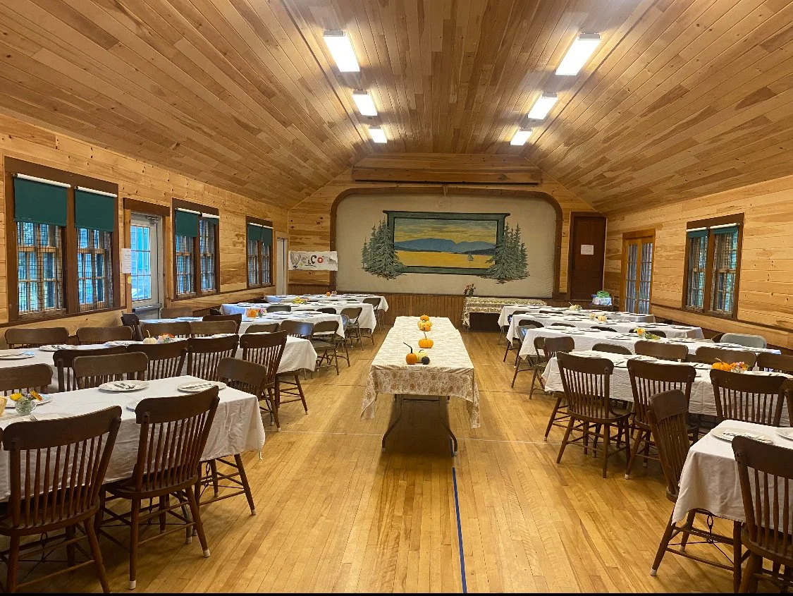 A large dining room with wooden walls and a vaulted ceiling, set up with tables covered in white tablecloths and surrounded by wooden chairs, decorated for a meal or event, with pumpkins and small decorations on the tables.