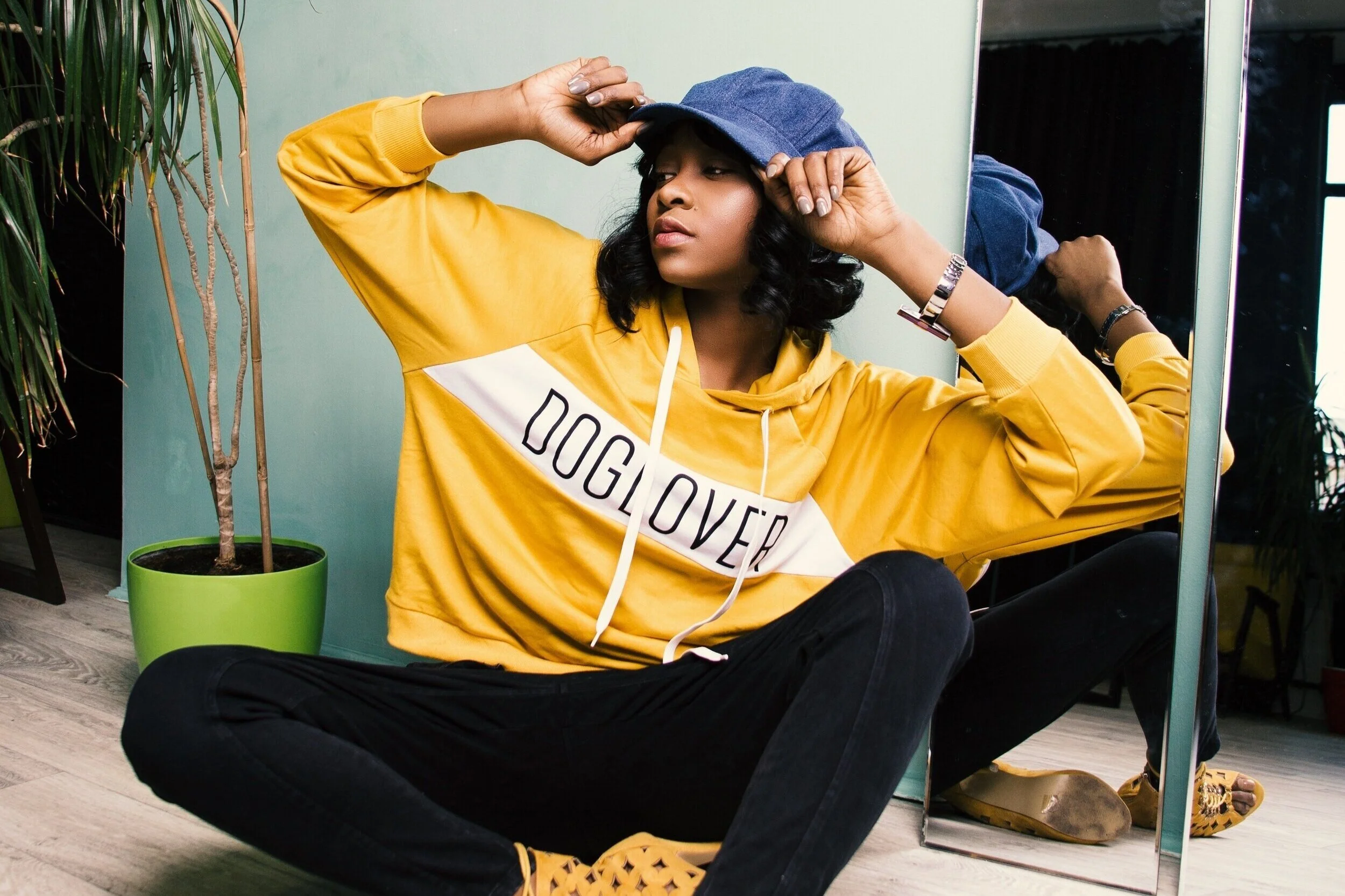 Woman sitting on the floor, adjusting a blue cap, wearing a yellow hoodie with 'DOG LOVER' written on it, black pants, and yellow shoes with a leopard print in front of a mirror, next to a green potted plant.