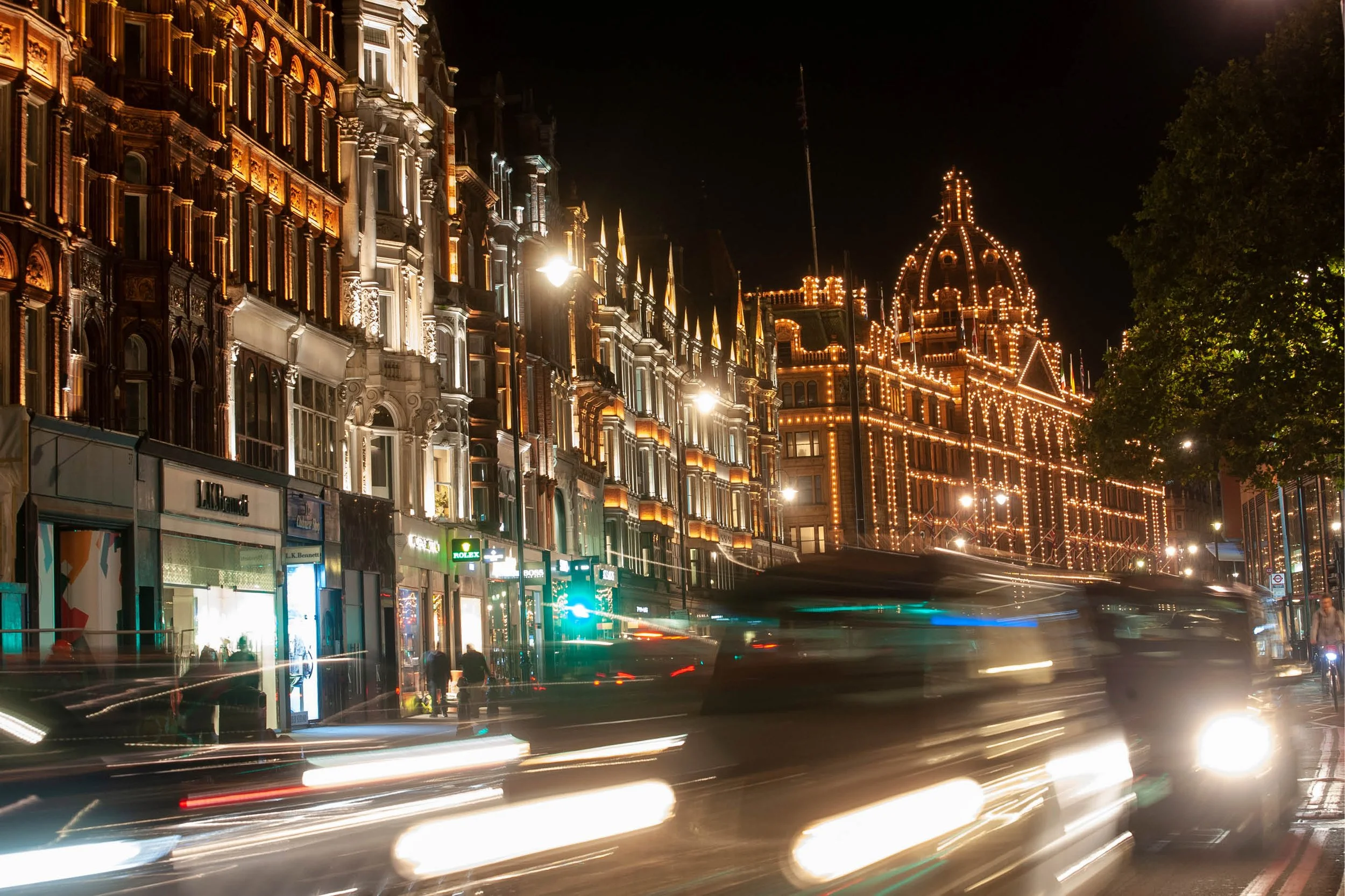 Nighttime city street with historic buildings decorated with string lights, blurred moving vehicles, and pedestrians on the sidewalk.