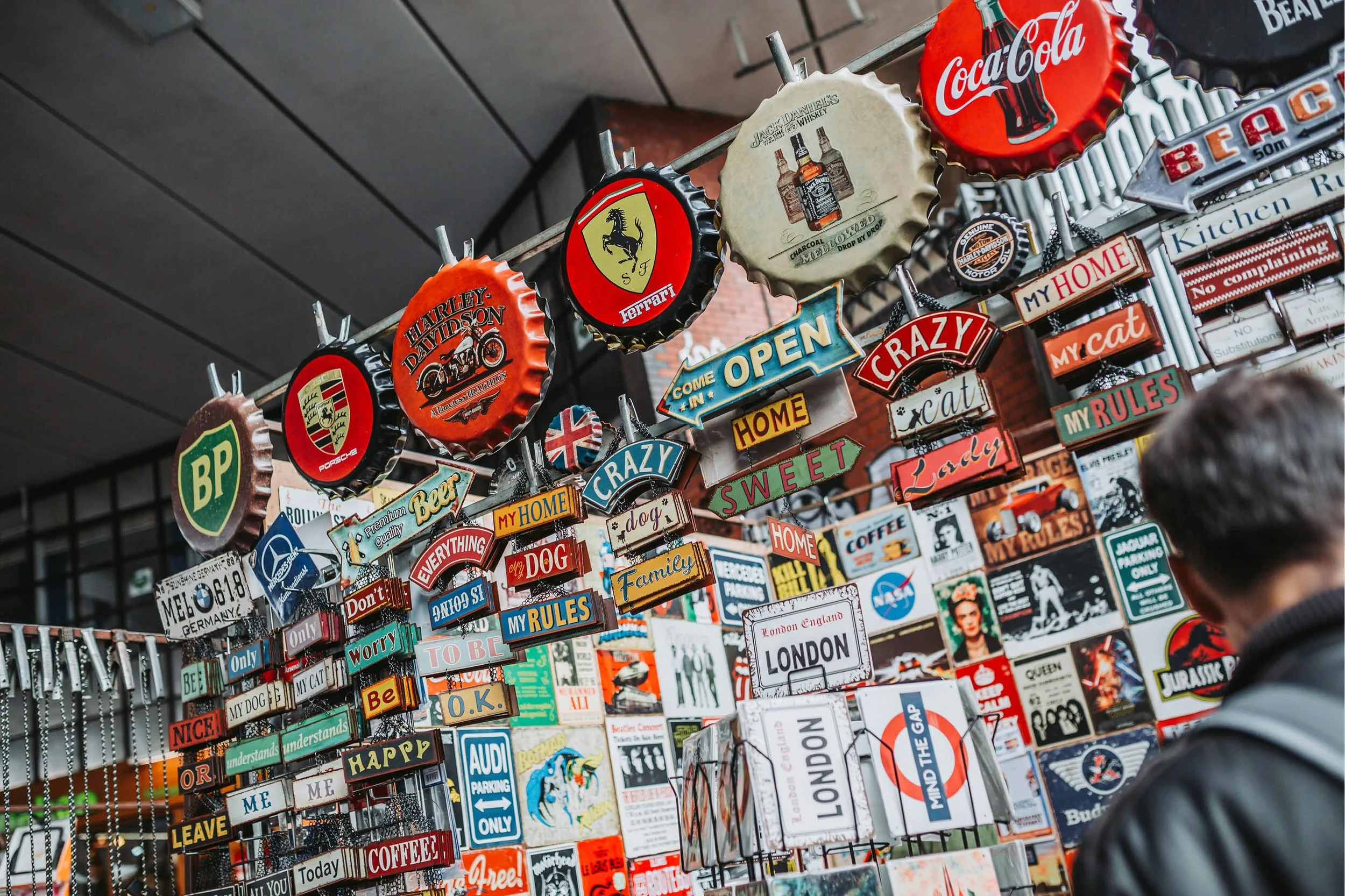 Display of colorful souvenirs including signs and bottle caps with logos of brands like Coca-Cola, Ferrari, Harley Davidson, Porsche, and Mercedes-Benz, as well as signs with text such as "Come in, We are open," "My Rules," "Dog," "Family," "London," and "Nice!" in an indoor marketplace setting.