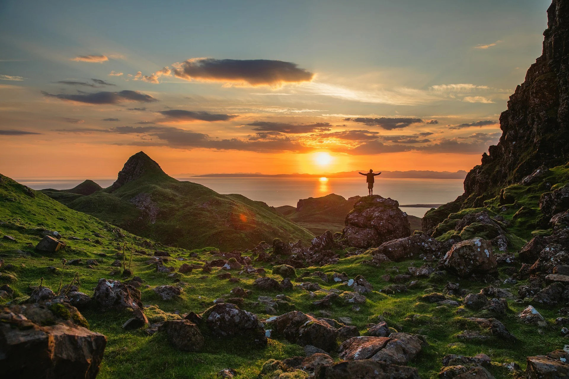 A man standing in his power after coaching with arms outstretched on a rock at sunset, overlooking green hills, rocky terrain, and a body of water in the distance.