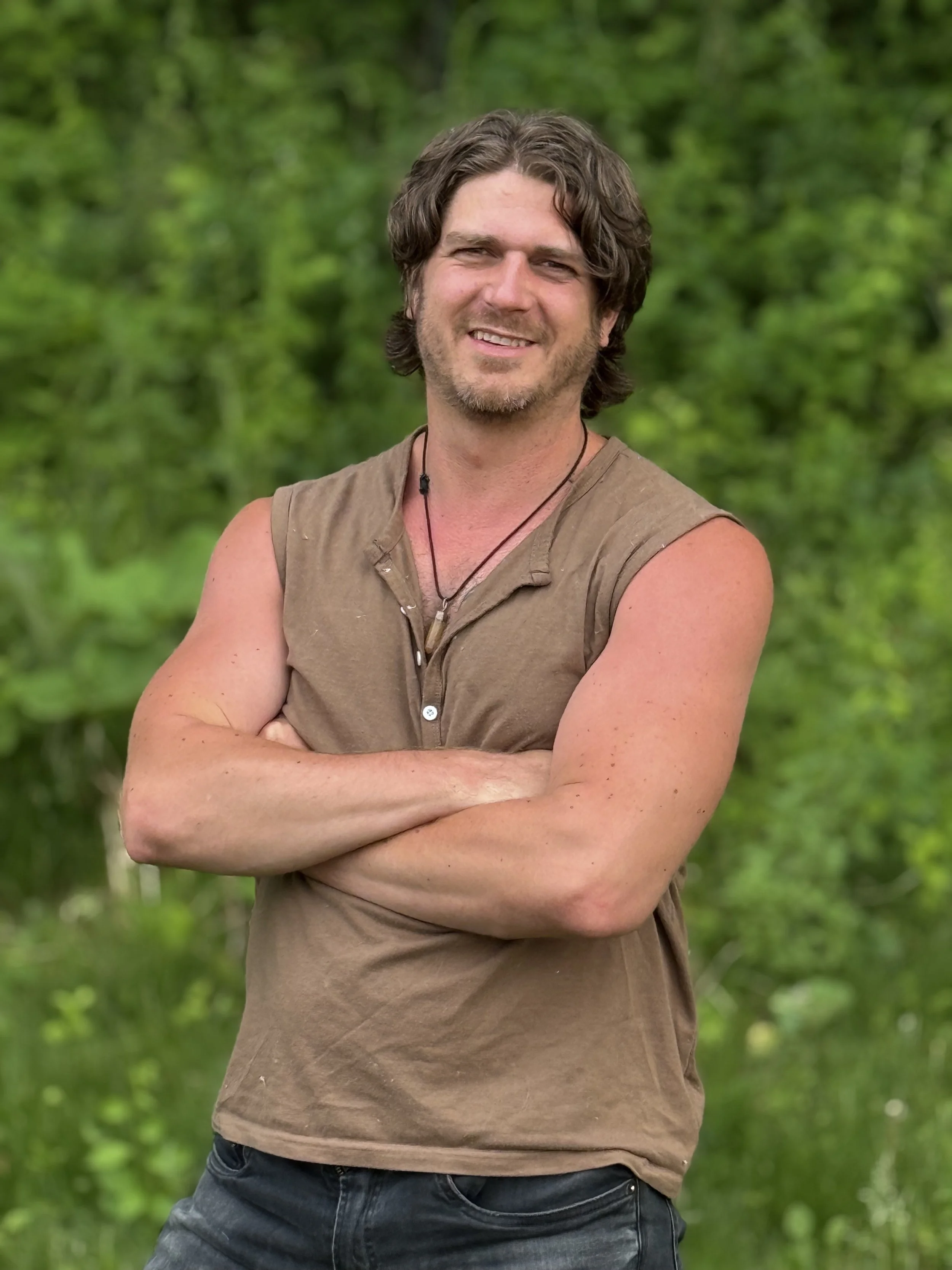 Greg Nordeng, Clear Mirror Coach for men is standing outdoors with arms crossed, smiling, wearing a sleeveless brown shirt and jeans, with a green blurred forest background.