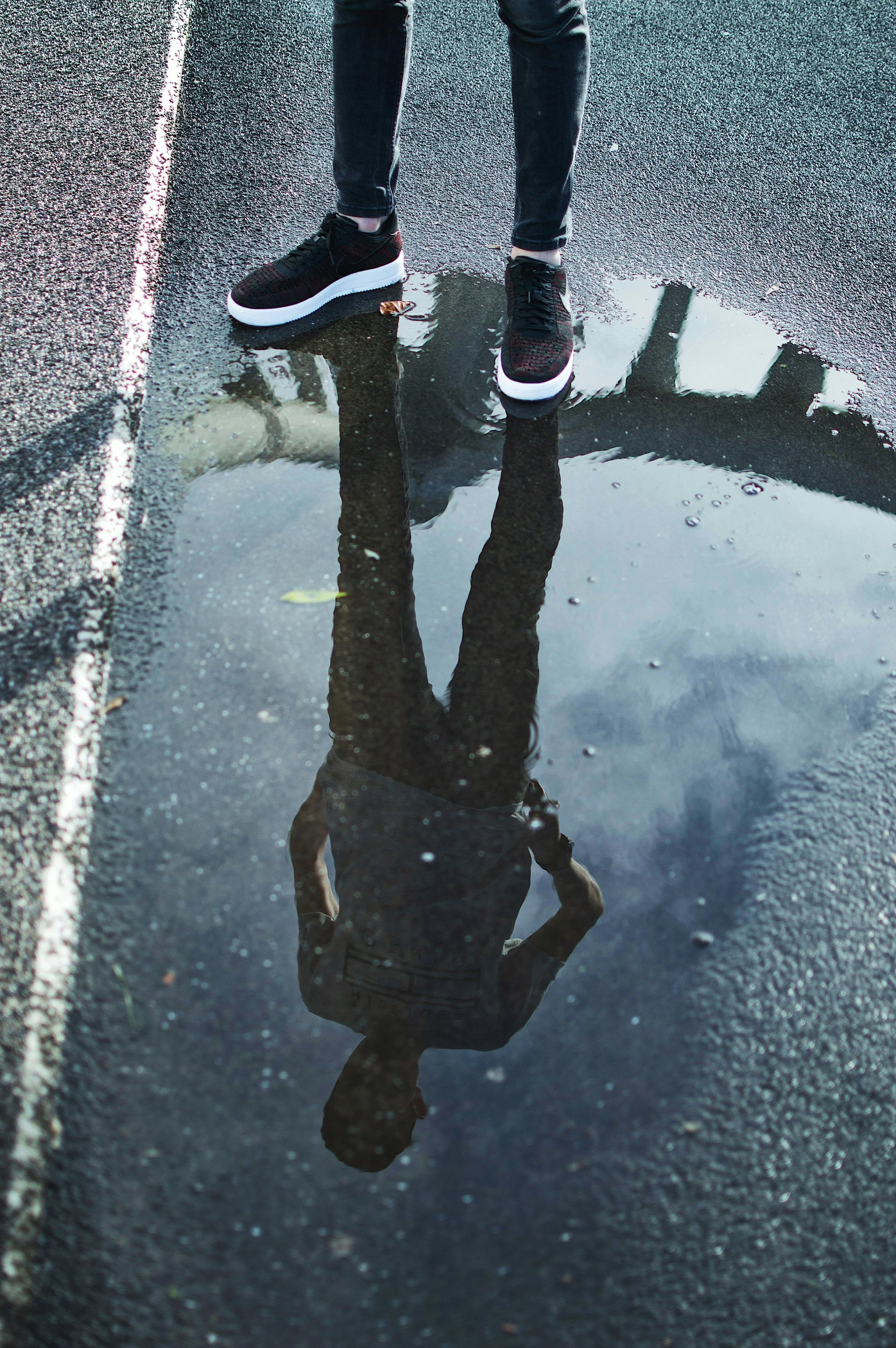 Person standing on wet pavement with puddle, their reflection visible in the water, wearing black sneakers and dark pants.