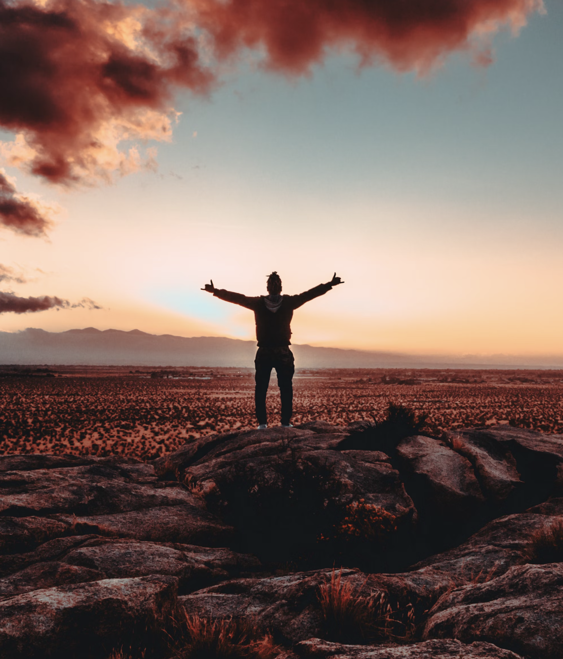 A person standing on rocks with arms outstretched, facing a desert landscape at sunset with colorful clouds in the sky.
