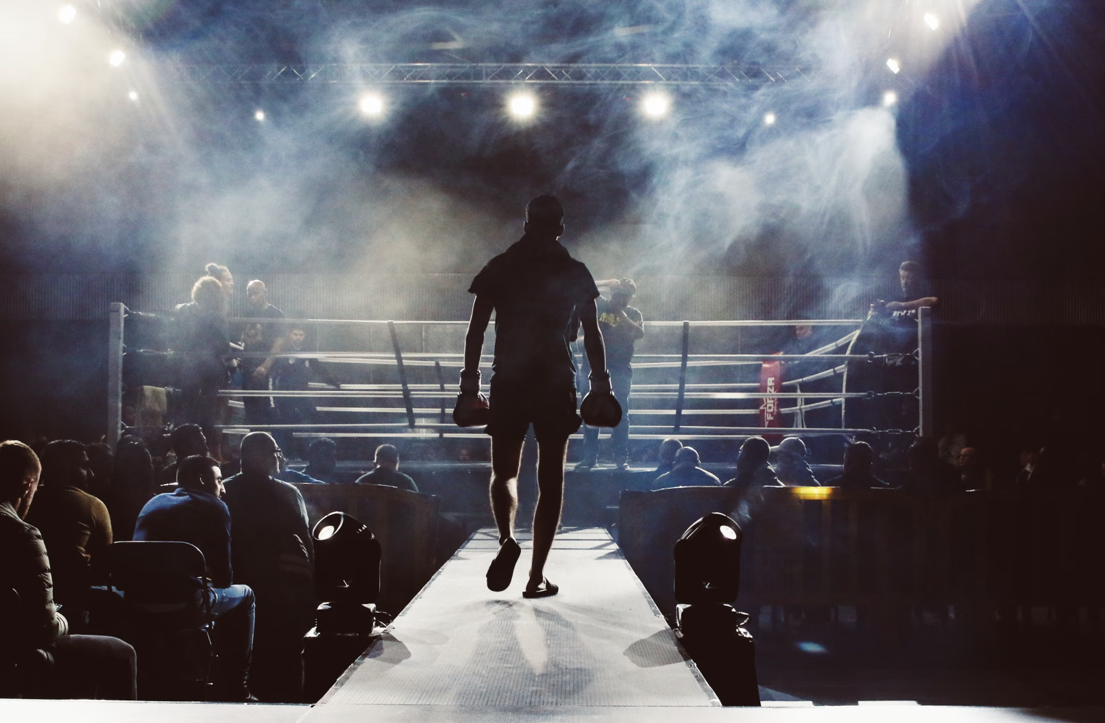 A silhouetted male boxer walking on a runway towards a boxing ring, surrounded by an audience with stage lights and smoke effects.