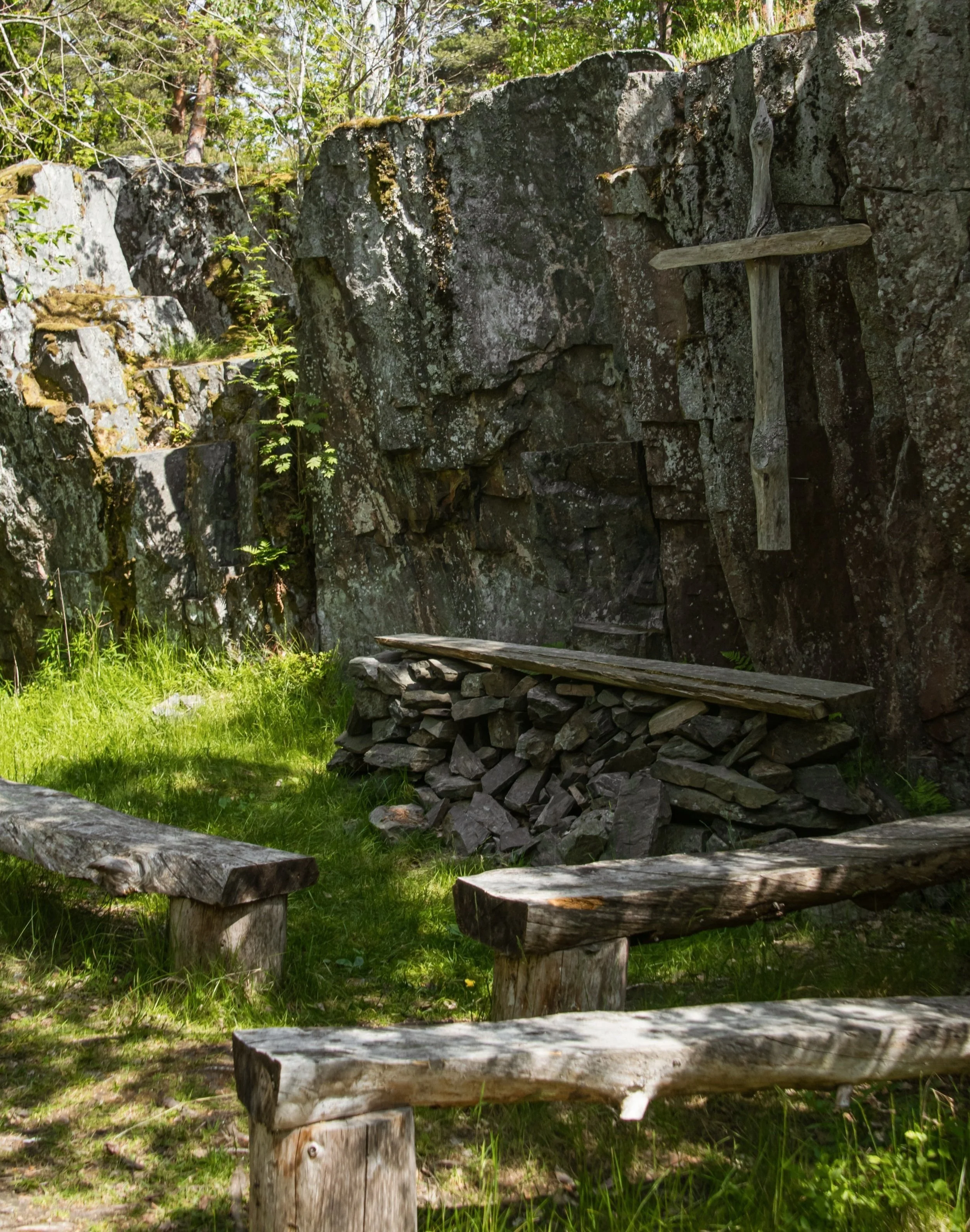 Outdoor scene with wooden benches and a stone wall, surrounded by greenery and trees, forest church at Vartiosaari, Helsinki, Finland