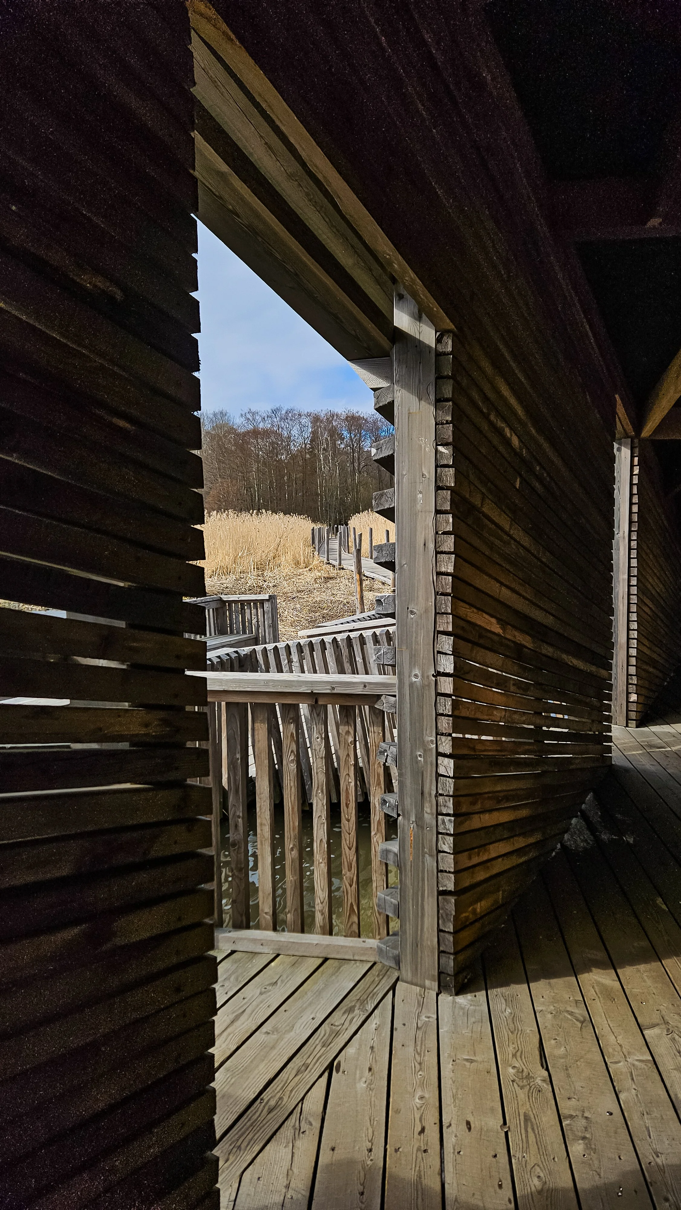 View through a wooden slatted wall and an opening shows a bridge railing, a wooded area, and tall grass in the distance under a partly cloudy sky, Lammassaari, Helsinki, Finland