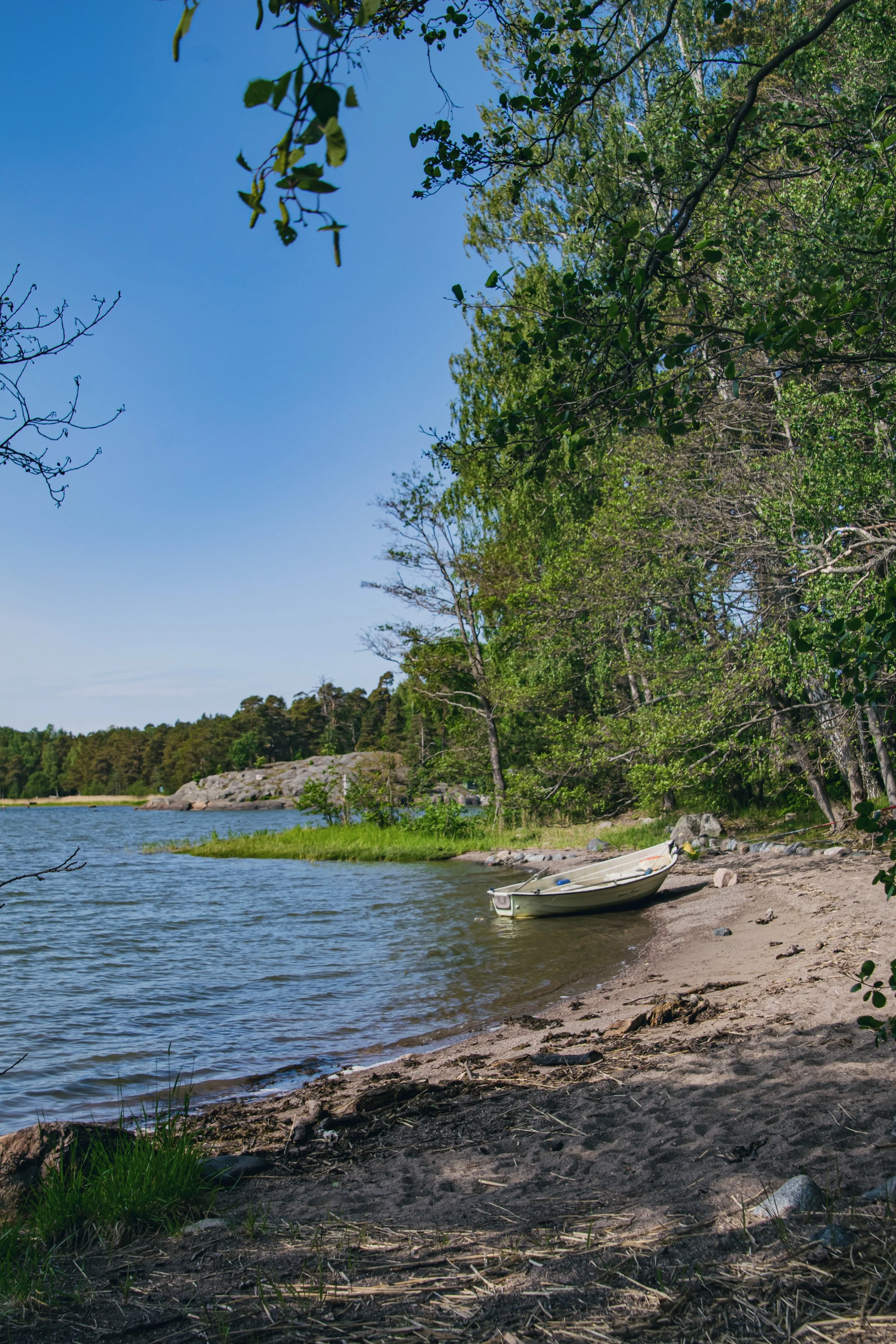 A small boat rests on a sandy shoreline near a lake, surrounded by trees with green leaves and rocks under a clear blue sky, Vartiosaari, Helsinki, Finland