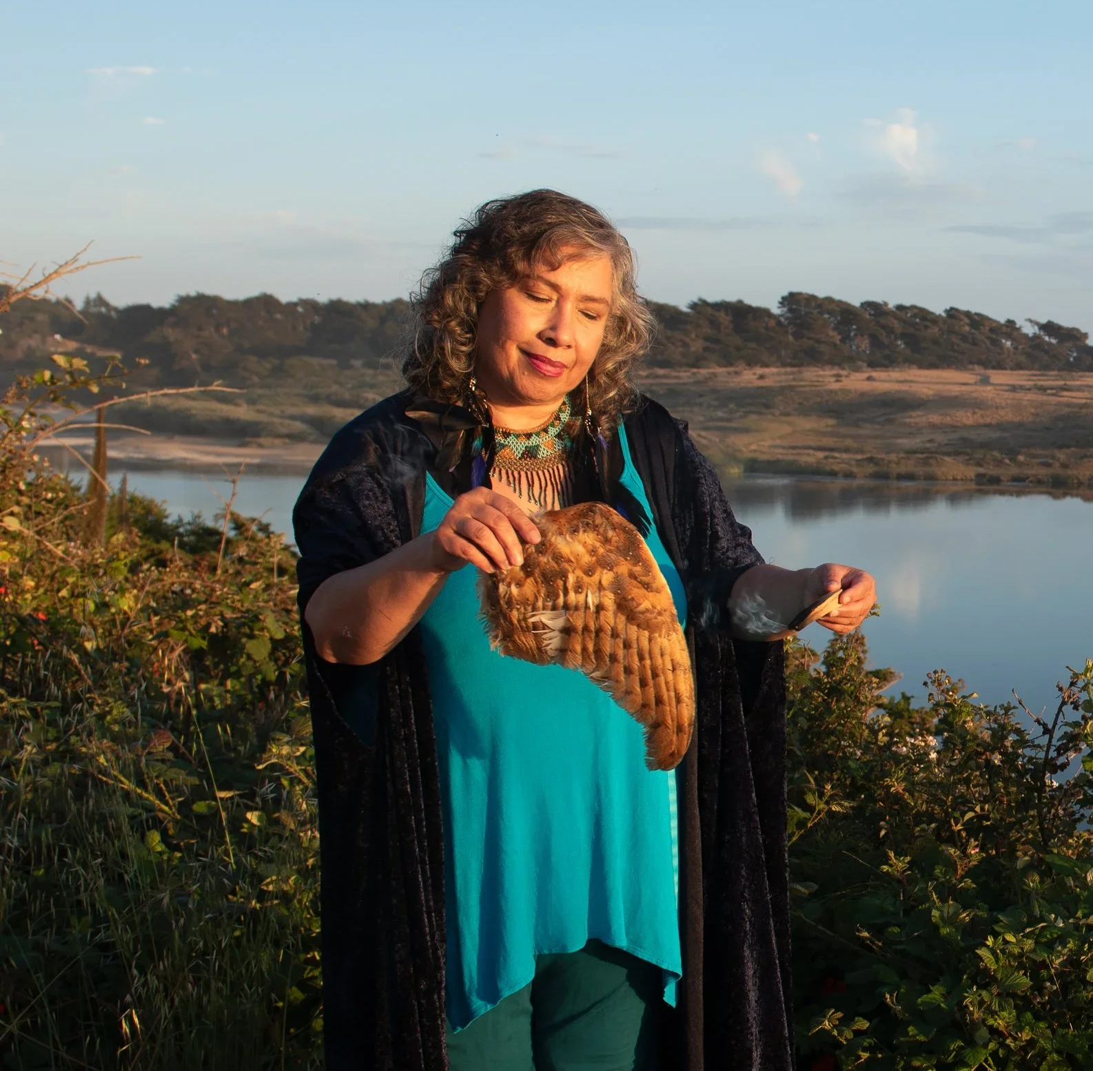 A woman stands outdoors near a body of water, holding a large owl on her arm, with a landscape of water and hills in the background during sunset.
