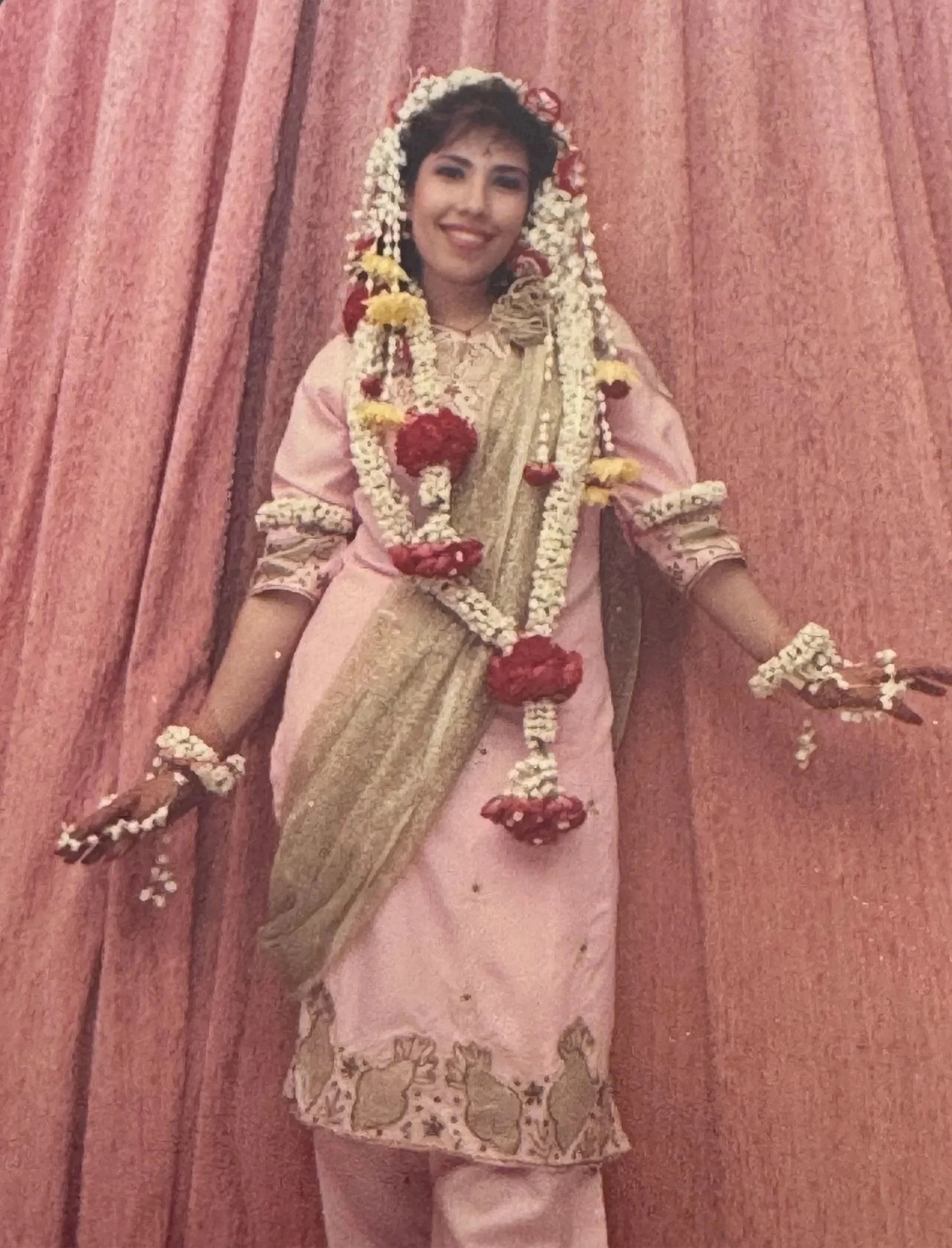 Veenaa Saynana Dolphin Heart Alchemy dressed in traditional South Asian attire, standing in front of a pink curtain. She is adorned with floral garlands and jewelry, and is smiling.
