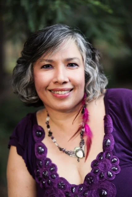 Veenaa Saynana Dolphin Heart Alchemy smiling outdoors, wearing a purple top with decorative stitching and a beaded necklace.