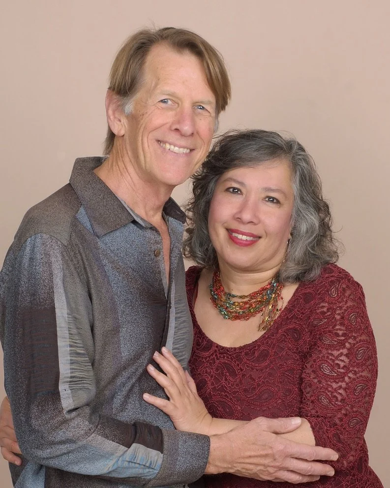 Veenaa Saynana Dolphin Heart Alchemy and her partner standing close together against a plain beige background, with the woman wearing a red lace top and the man in a multicolored button-up shirt.