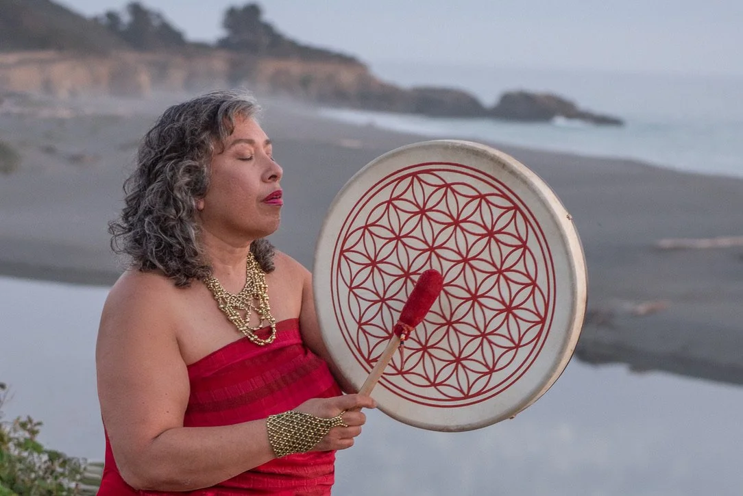 A woman in a red strapless dress with jewelry stands on a beach holding a decorated drum and a mallet with red fur, with the ocean and cliffs in the background.