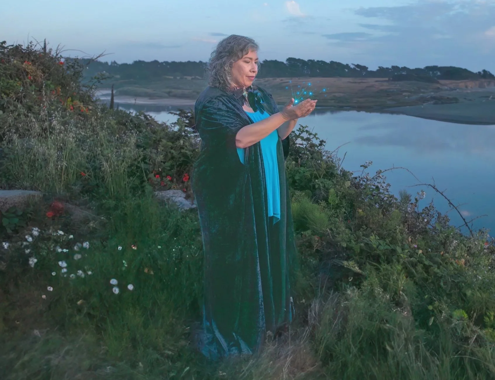 Veenaa Saynana Dolphin Heart Alchemy stands on a grassy riverbank at sunset, holding her hands in front of her and looking at a glowing, digital constellation of blue stars above her palms, with a lake and rolling hills in the background.