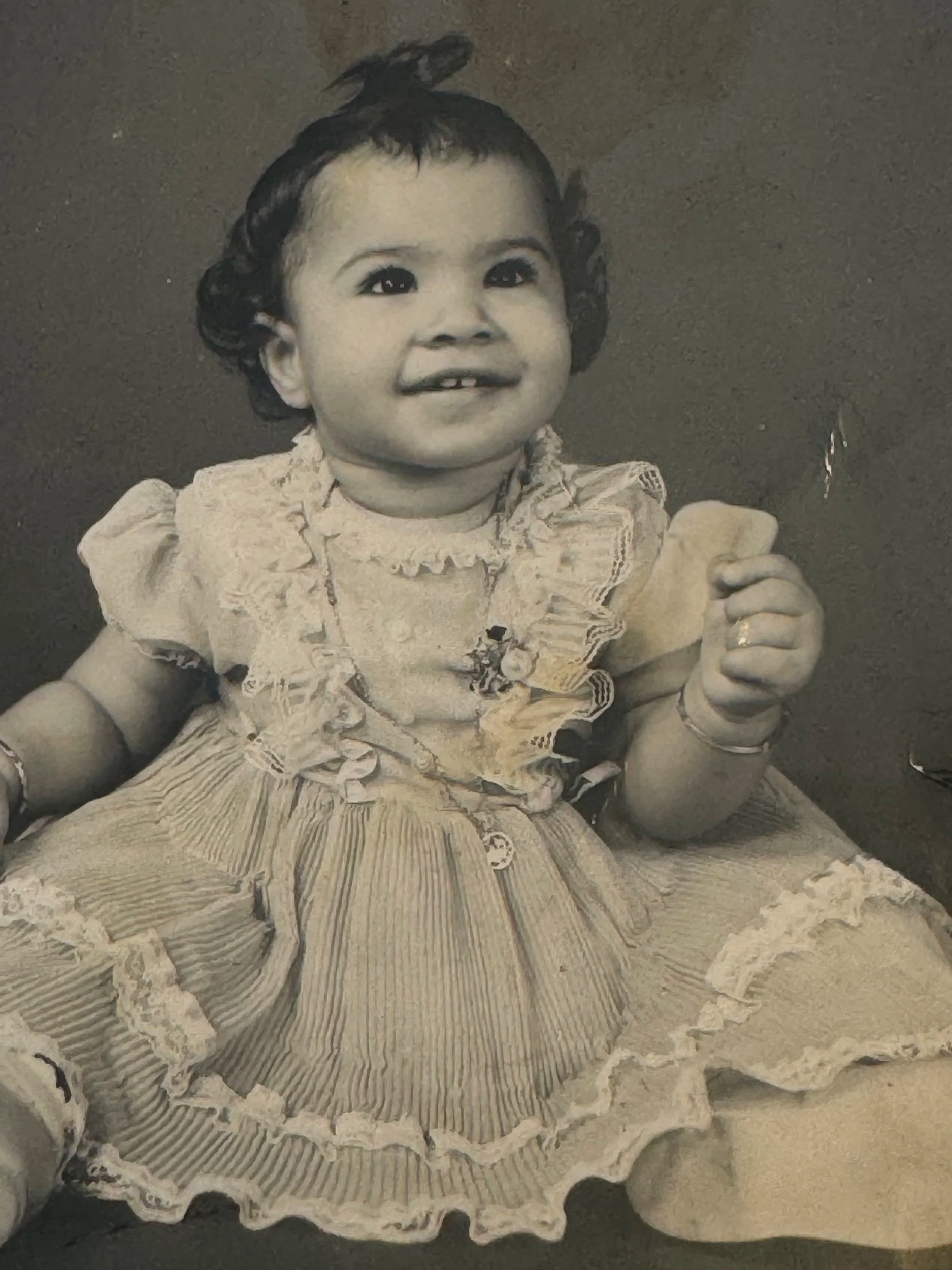 Black and white photo of a smiling young Veenaa Saynana Dolphin Heart Alchemy wearing a dress with ruffles and lace, against a plain dark background.