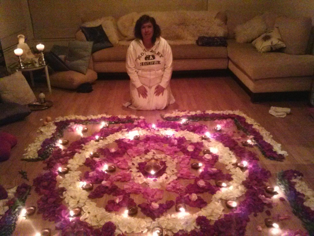 Veenaa Saynana Dolphin Heart Alchemy kneeling on the floor in a living room with a large floral rangoli decorated with candles and rose petals.