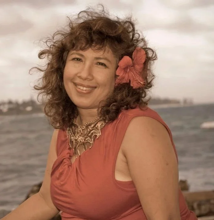 Veenaa Saynana Dolphin Heart Alchemy with curly hair and a flower hair accessory smiling outdoors near water on a cloudy day.