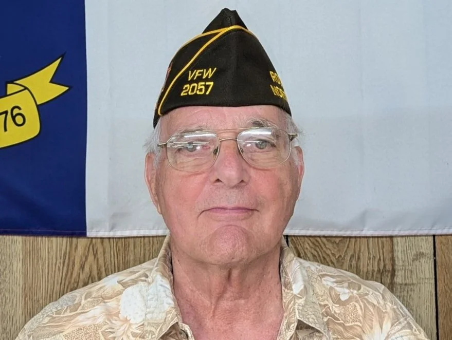 An elderly man wearing glasses and a black VFW military cap with yellow text, standing in front of a flag with a blue background and yellow emblem.