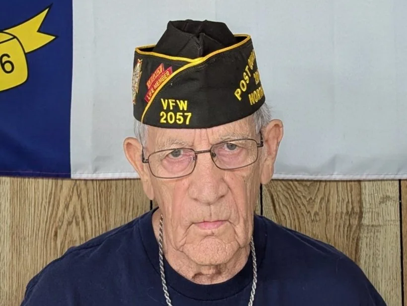 An elderly man wearing glasses and a black veteran's hat with yellow lettering, sitting in front of a flag and wood-paneled wall.