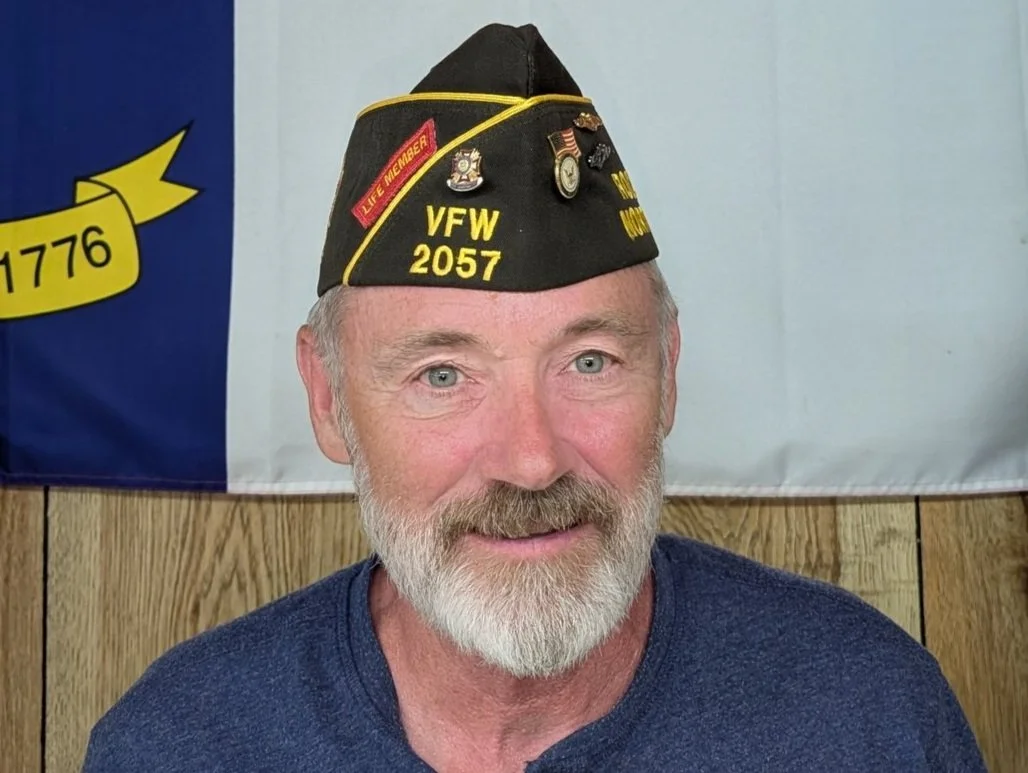 A smiling man with a gray beard wearing a black cap with military pins and patches, and a navy blue shirt, standing in front of a flag with a blue and yellow banner.
