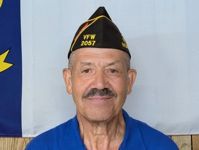 A smiling elderly man wearing a black veterans of foreign wars hat with yellow lettering, a blue shirt, and a background with a blue and white design.