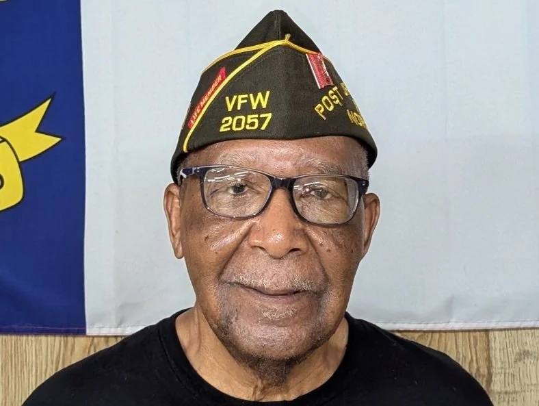 A senior man wearing glasses and a black VFW hat that reads 'VFW 2057 POST' standing indoors in front of a wall with a flag partially visible on the left.